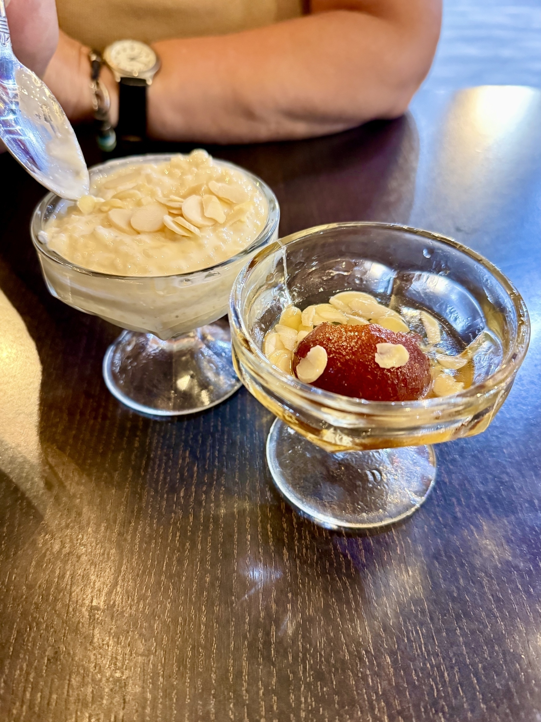 Two glass cups of dessert on a wooden table. The left cup contains rice pudding topped with almond slices, and a spoon is scooping some out. The right cup holds a gulab jamun in syrup, also garnished with almond slices. A person wearing a watch with a black strap is partially visible in the background.