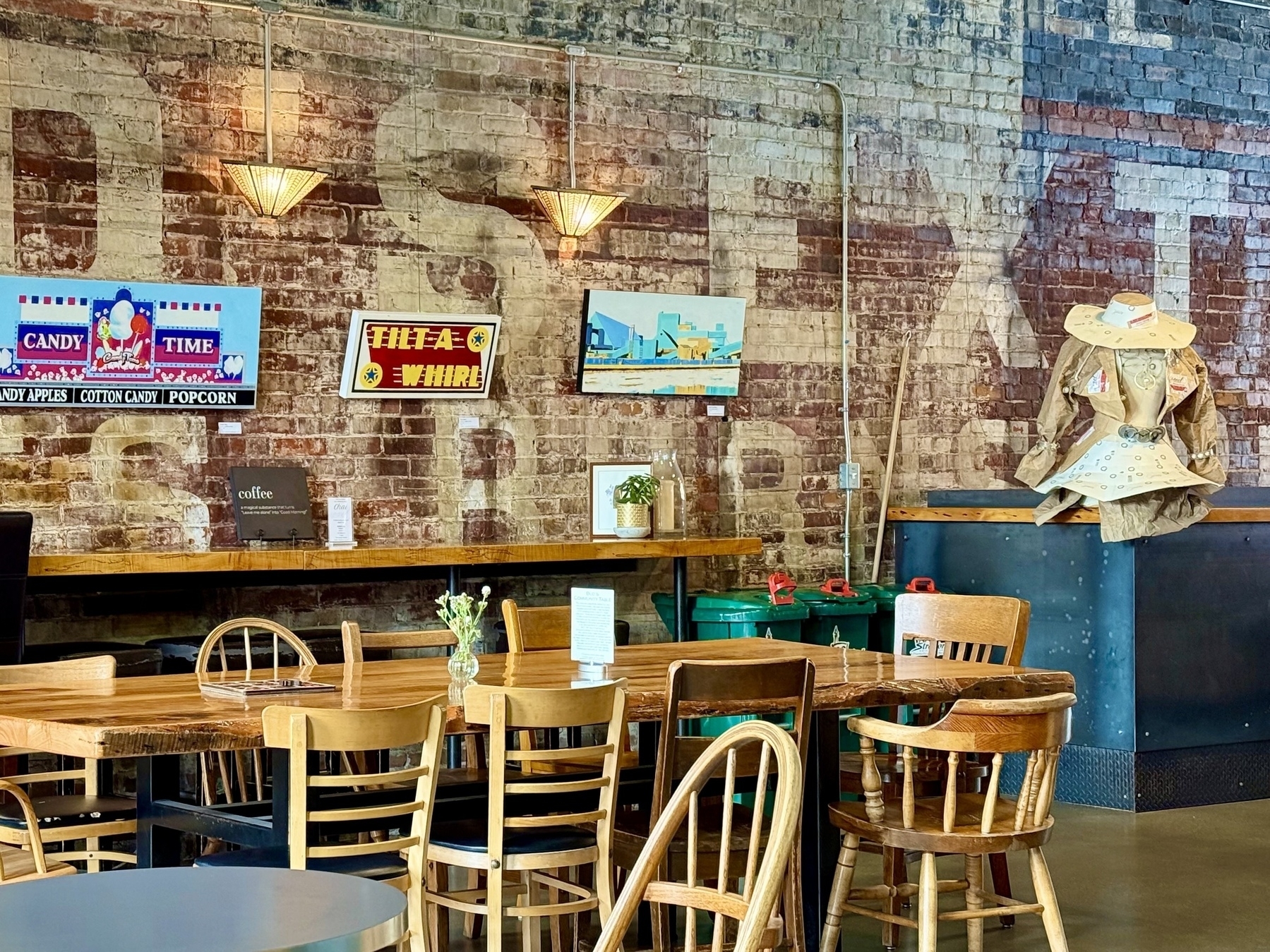 A cozy interior of a cafe with a rustic brick wall adorned with vintage-style signs, one reading "CANDY TIME" and another "TILT-A-WHIRL". There are a few framed artworks on the wall. Wooden tables and chairs are arranged for seating, and a small vase with flowers is placed on a table. On the right, a coat stand displays what appears to be a decorated coat and hat. Industrial-style pendant lights hang above, casting a warm glow over the space.
