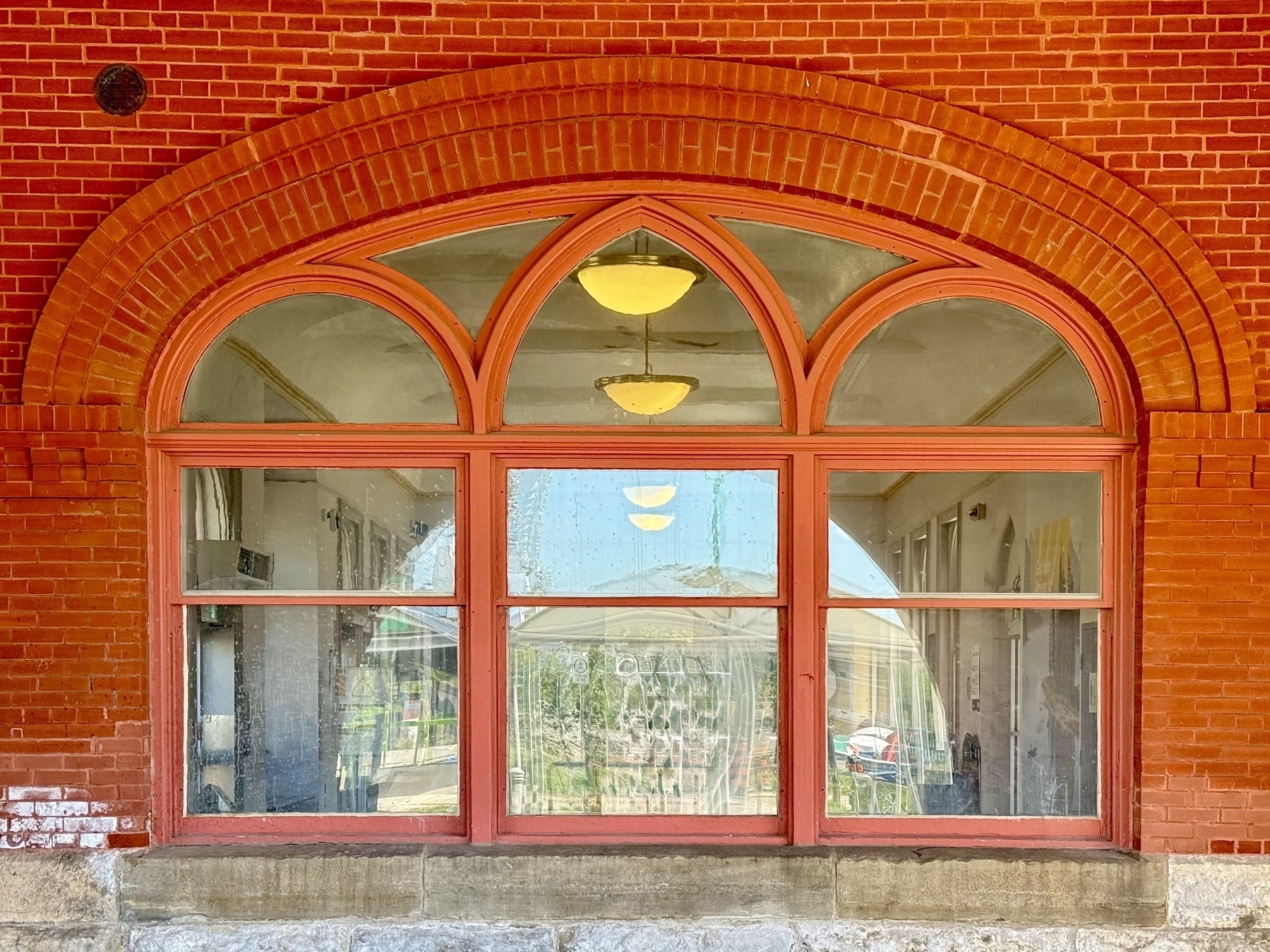 A red brick wall featuring an arched window with multiple small panes, framed in a slightly darker shade of red. The window reveals a corridor inside, where two hanging light fixtures are visible. The window's reflective surface partially shows the outdoor surroundings. The architectural design showcases a classic style with an arch above the window.
