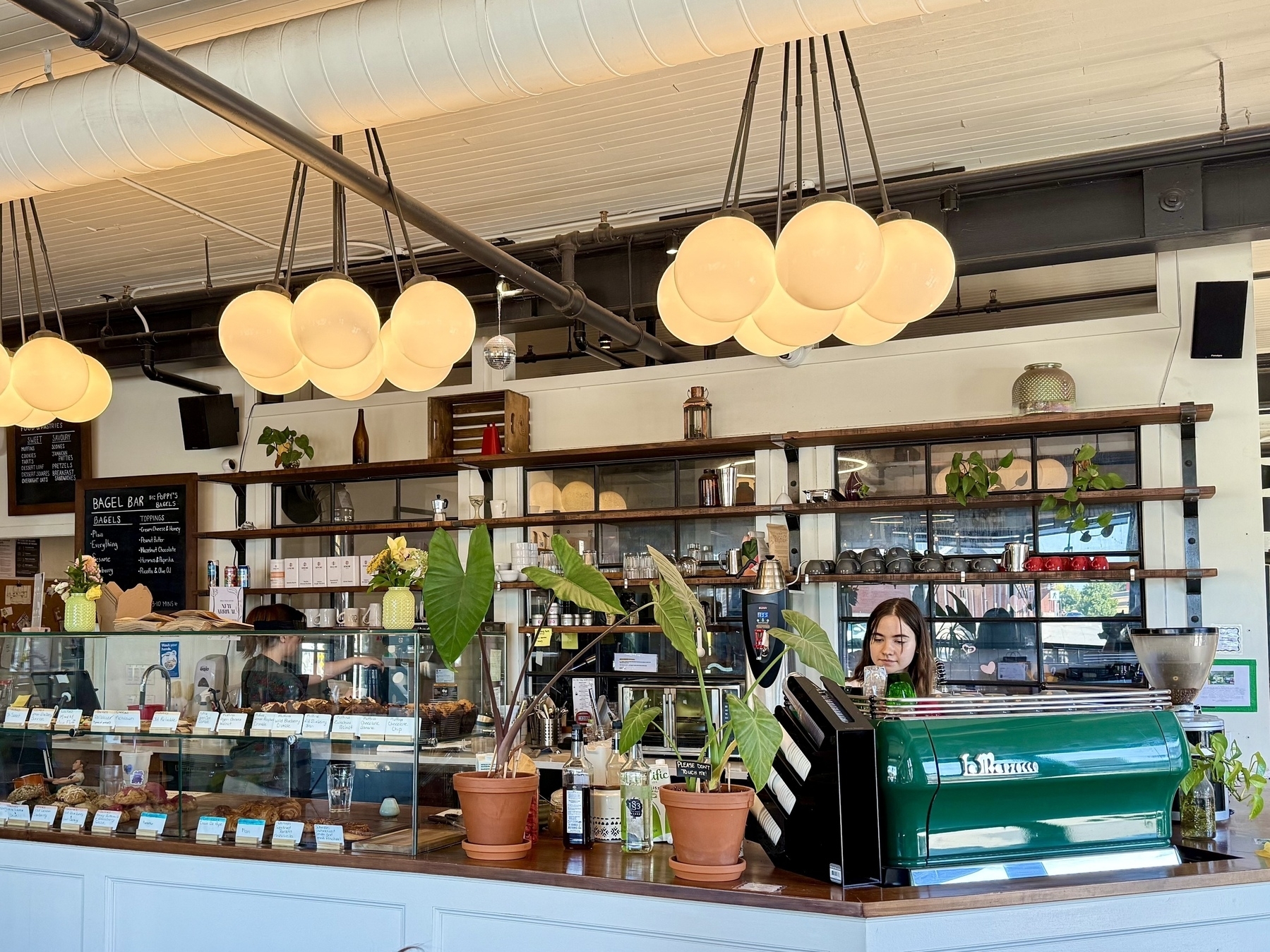 A cozy café interior with a long counter displaying various baked goods and pastries. The counter is topped with large green plants in pots. Above, three modern light fixtures with round bulbs hang from the ceiling. A person stands behind an espresso machine. The background features shelves with plants, cups, and small decor items. A chalkboard menu listing bagel options is visible on the left. The overall atmosphere is warm and inviting.