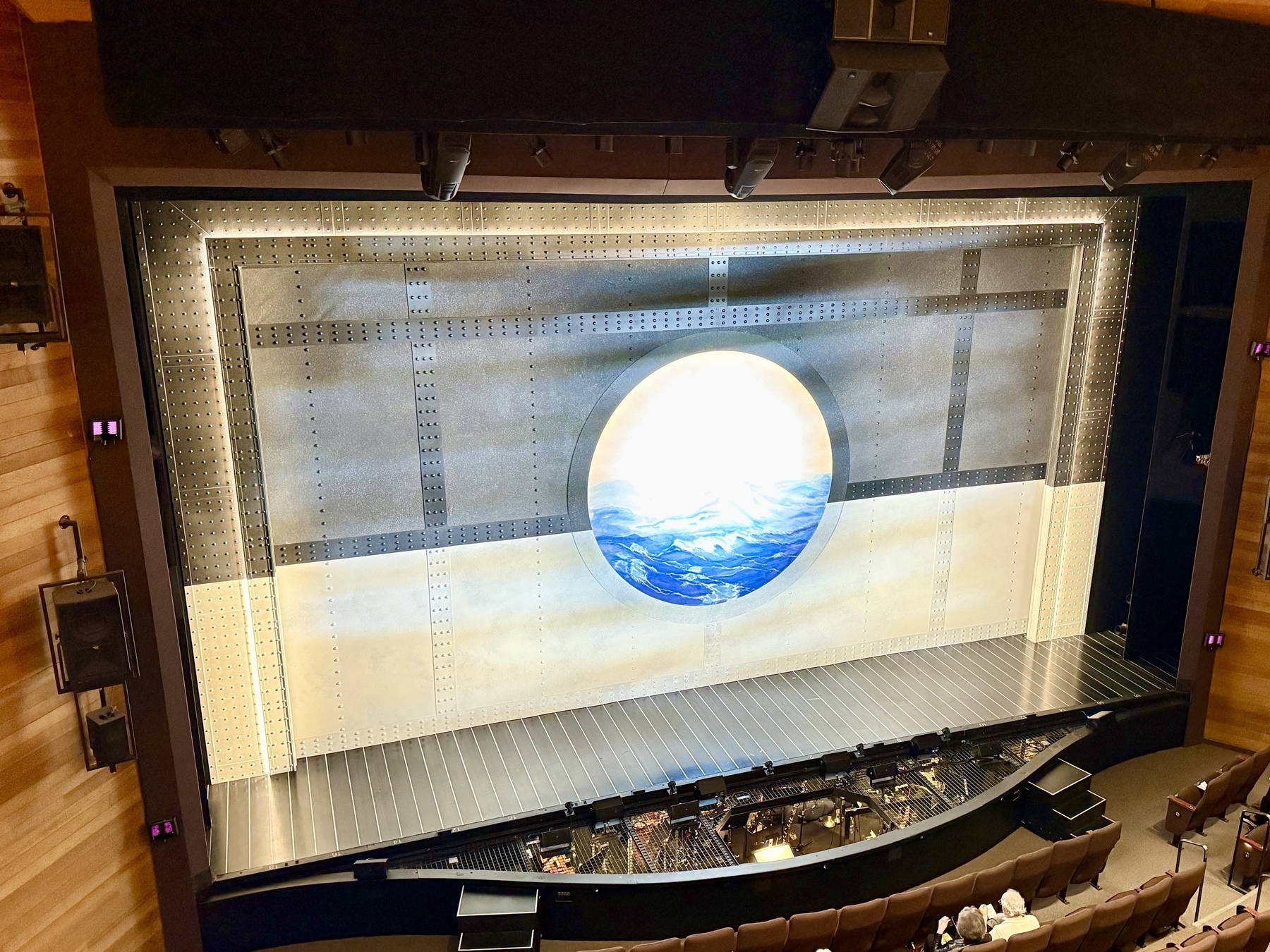 A theater stage set with a metallic, industrial design featuring a large circular window at the center displaying an image of mountains and a cloudy sky. The stage itself is dark with seating in the foreground, where a few audience members are visible, suggesting a view from a mezzanine or balcony.