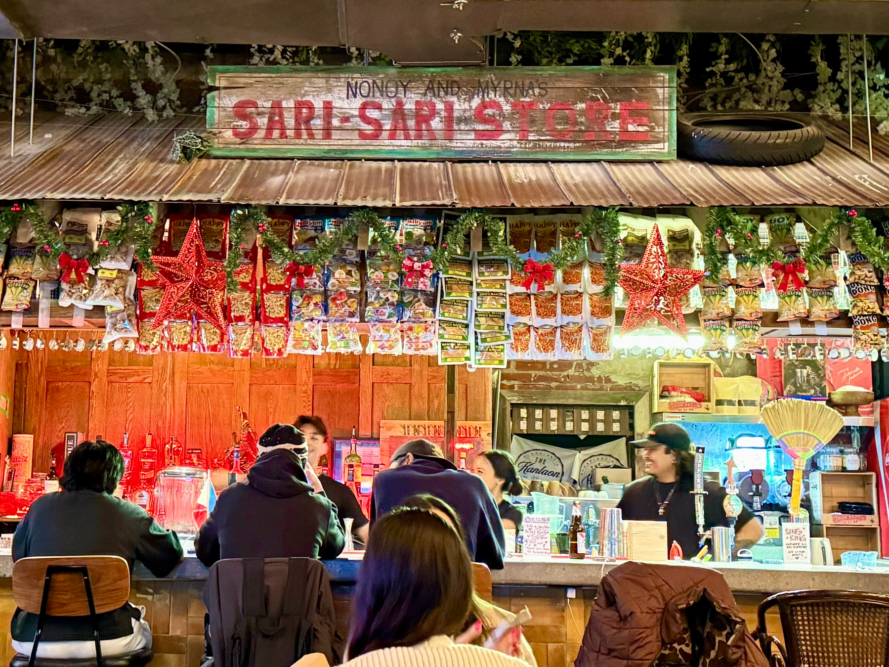 The image shows a lively scene at a "Sari-Sari" store-themed bar. The bar is decorated with colorful Christmas ornaments, wreaths, and red star lanterns. Various snacks are displayed hanging above the counter. Several people are seated at the bar, while attendants engage with customers. The decor includes traditional Filipino items like a broom (walis tingting) and a vintage style sign that reads "Nonoy and Myrna's Sari-Sari Store."