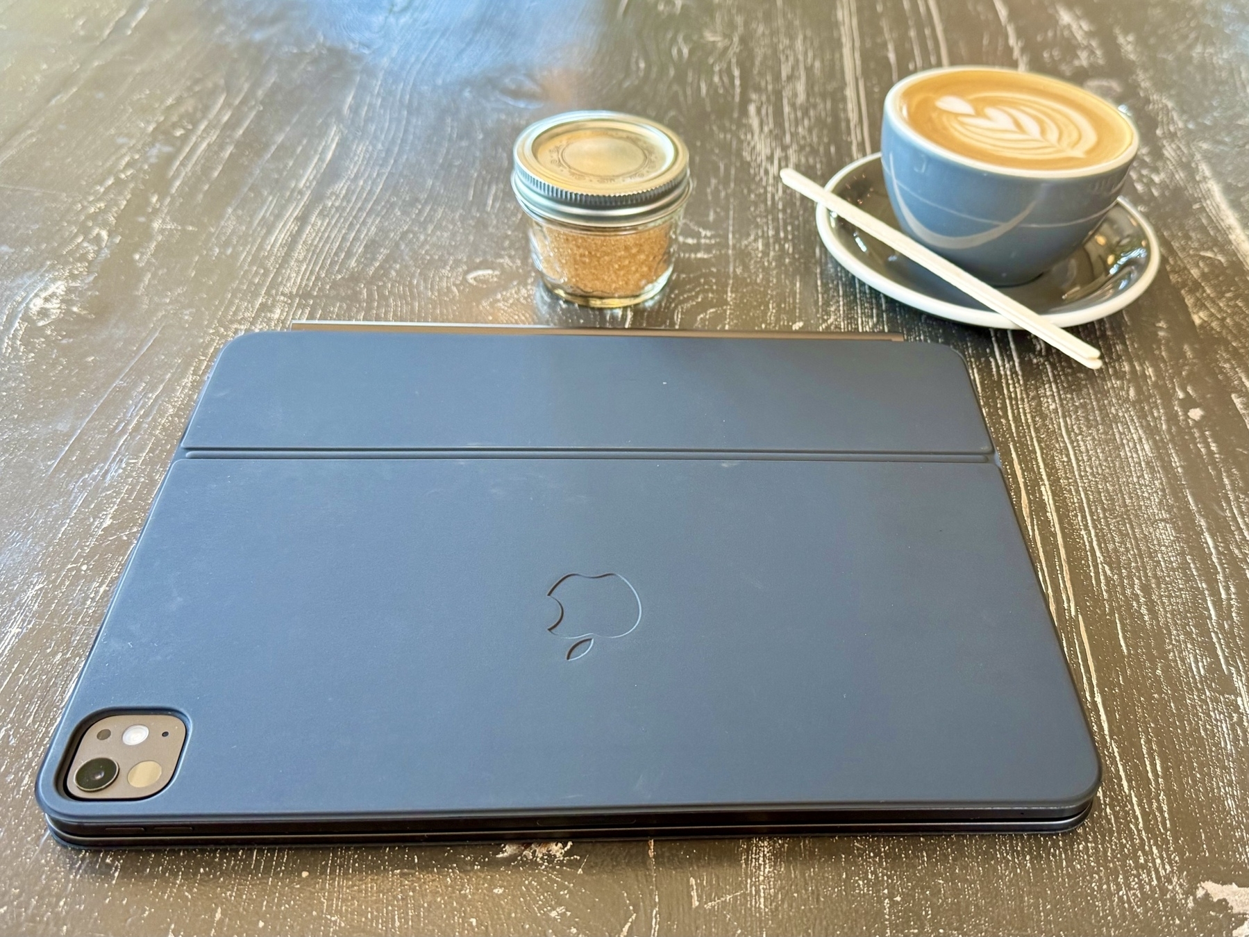 A closed black iPad with a visible camera is on a wooden table. Next to it is a small jar of brown sugar and a cup of cappuccino with latte art, placed on a saucer with a spoon.