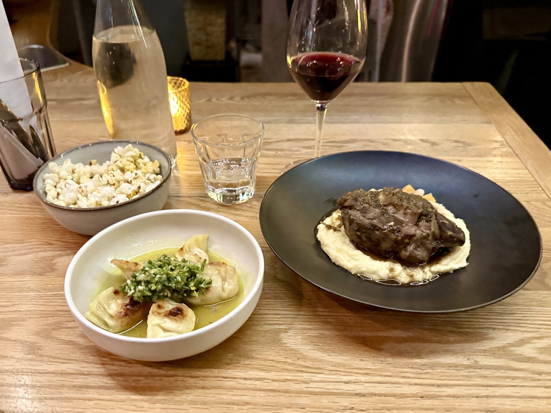 A wooden table set with several dishes and drinks. On the left, a bowl of popcorn. In the center, a dish with dumplings topped with green garnish in a light sauce. On the right, a plate with a piece of meat on a bed of mashed potatoes or similar puree. A glass of red wine and a clear bottle of water also sit on the table. In the background, there is a softly lit ambiance with a candle.