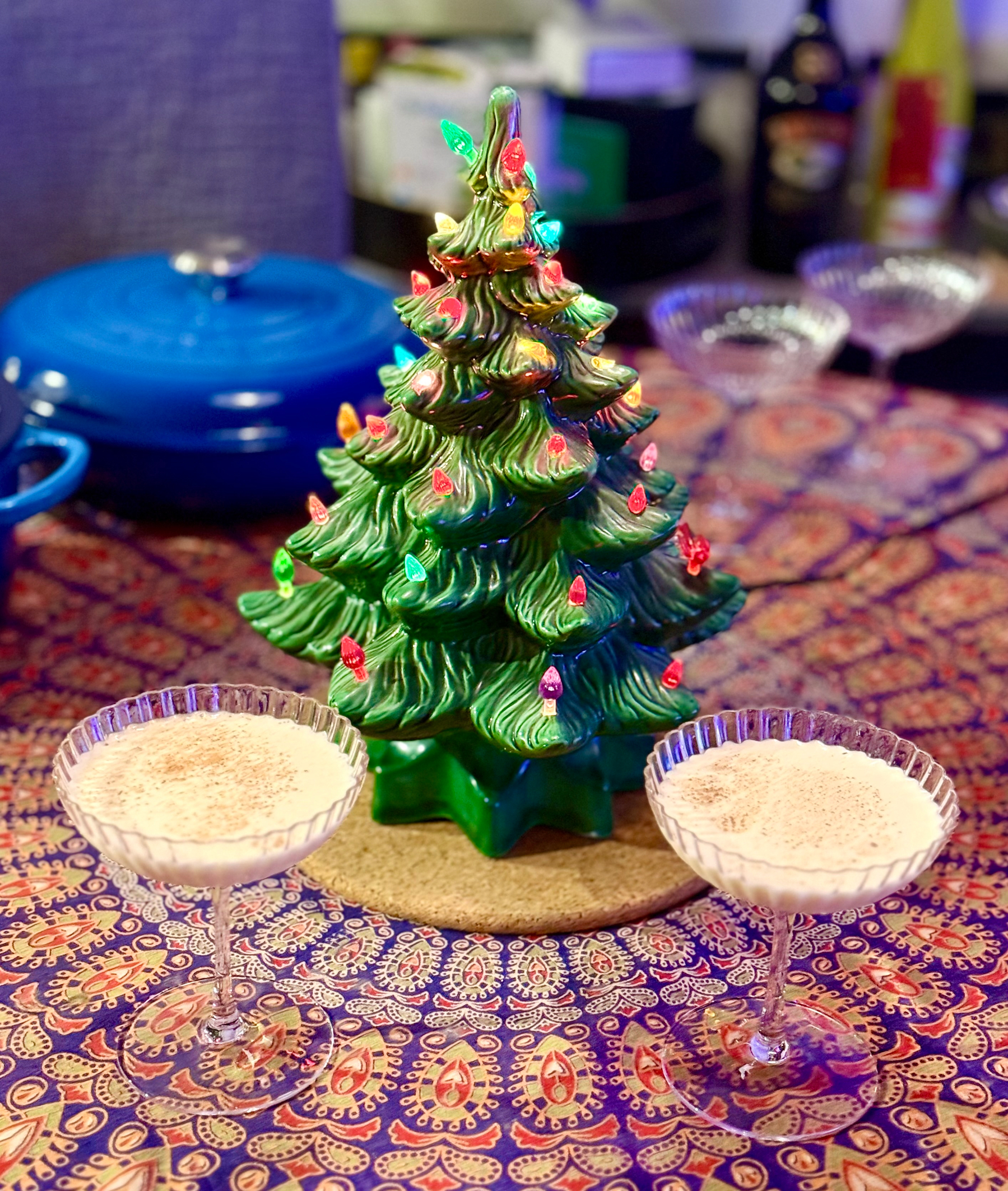 A small ceramic Christmas tree with colorful lights is centered on a table covered with a patterned cloth. Two glasses of creamy drinks are placed symmetrically on either side of the tree. A blue pot and some bottles are blurred in the background.