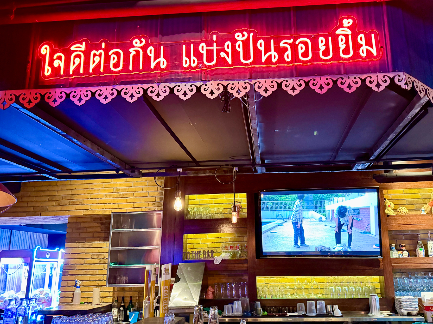 A neon sign with Thai script hangs above a bar with a television screen showing a scene from a Thai zombie series