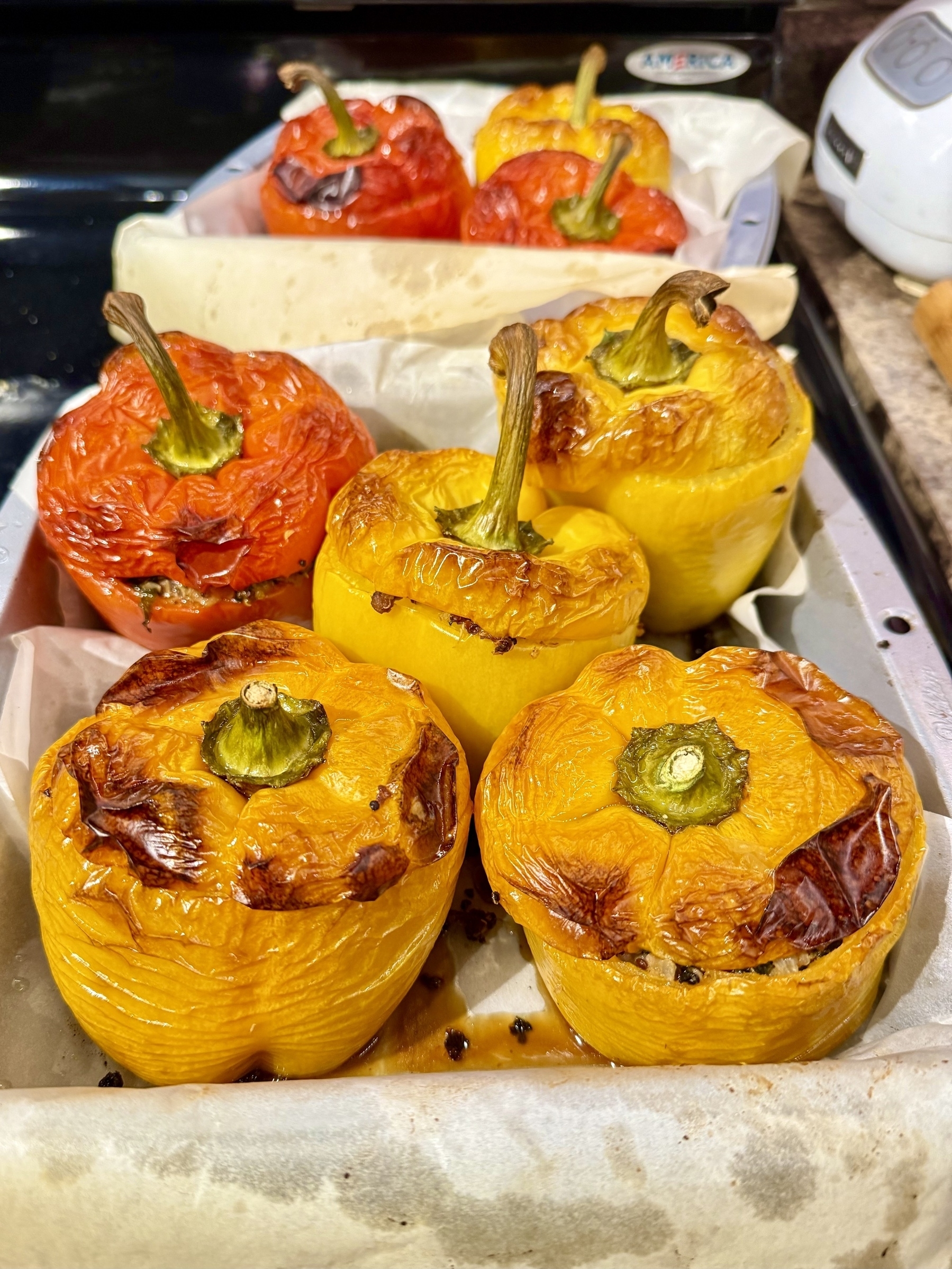Roasted yellow and red bell peppers placed in a baking dish lined with parchment paper. The peppers have a slightly blistered skin, indicating they have been cooked. They are stuffed with a visible filling under the pepper tops. The background shows parts of a kitchen counter and appliances.