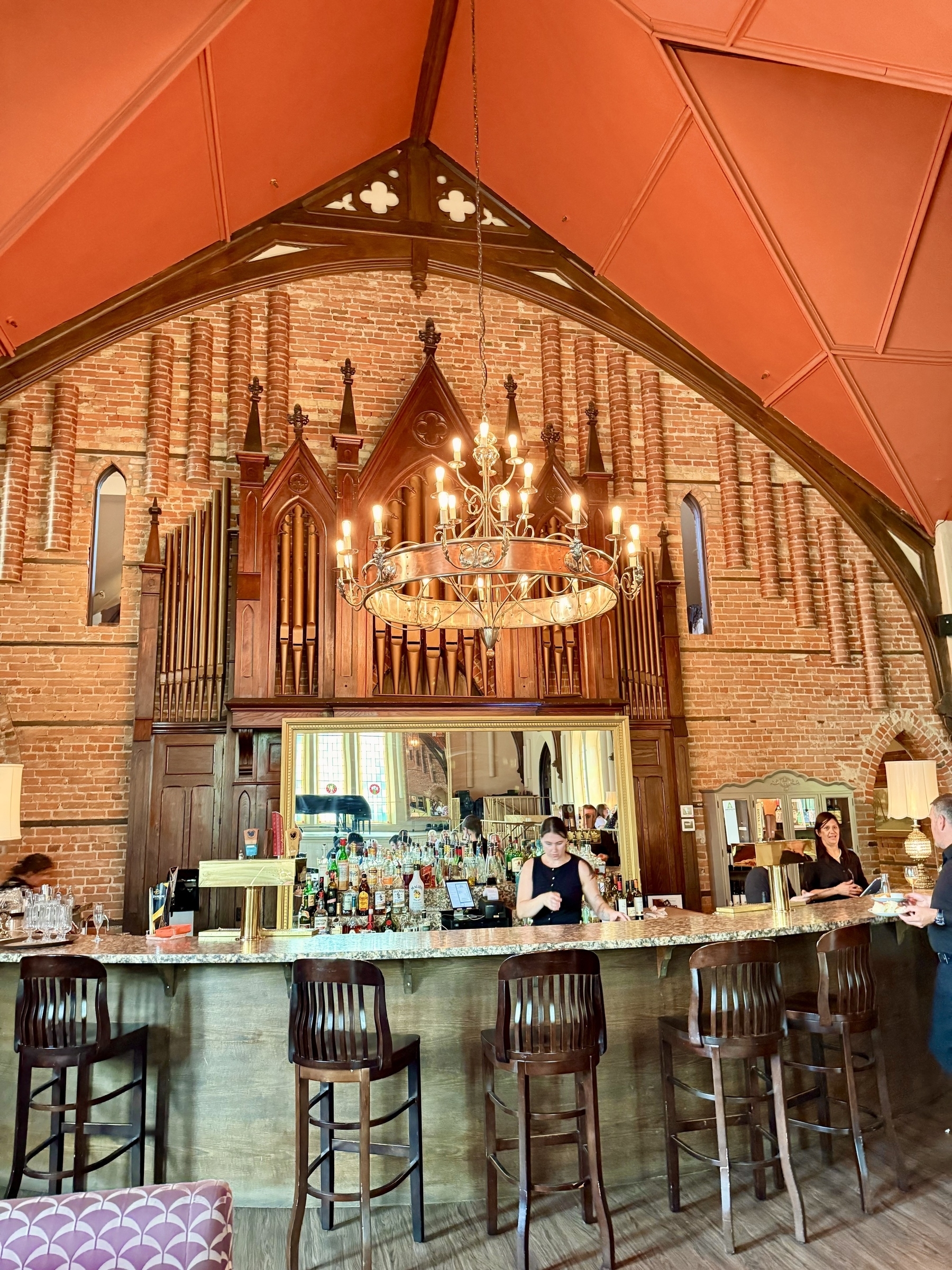 An ornate bar set inside a space designed like a cathedral, featuring a high, red-vaulted ceiling and brick walls with Gothic architectural elements. The bar is made of wood with a marble countertop and is stocked with various bottles. A large chandelier hangs overhead. Behind the bar, a large mirror reflects the room's interior. Several bar stools are placed in front, with one person working behind the bar and a couple of patrons standing nearby.