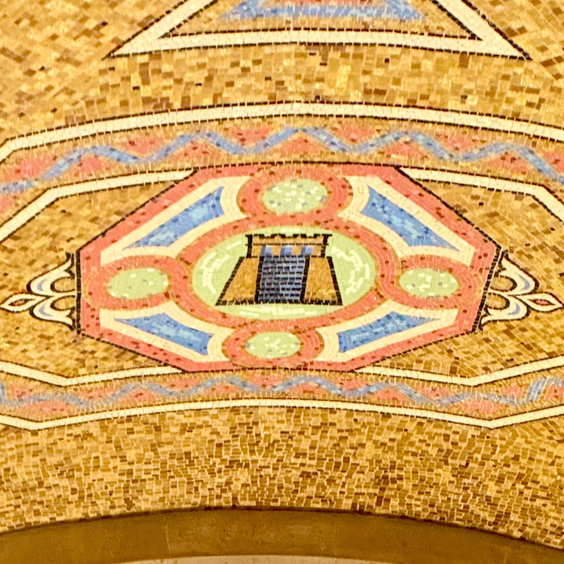 Intricate mosaic ceiling art featuring a central design with a Mayan? temple structure in blue tiles, surrounded by a hexagonal pattern. The surrounding colors include pink, blue, green, and gold, with additional geometric detailing. The background is composed of numerous small, gold-colored tiles.