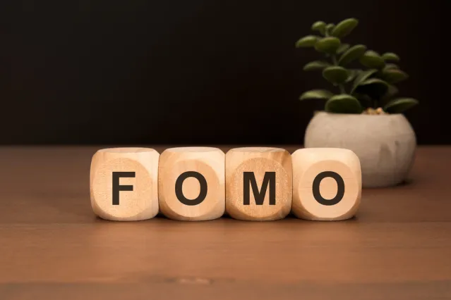 Wooden blocks spelling FOMO are placed next to a small potted plant on a wooden surface.