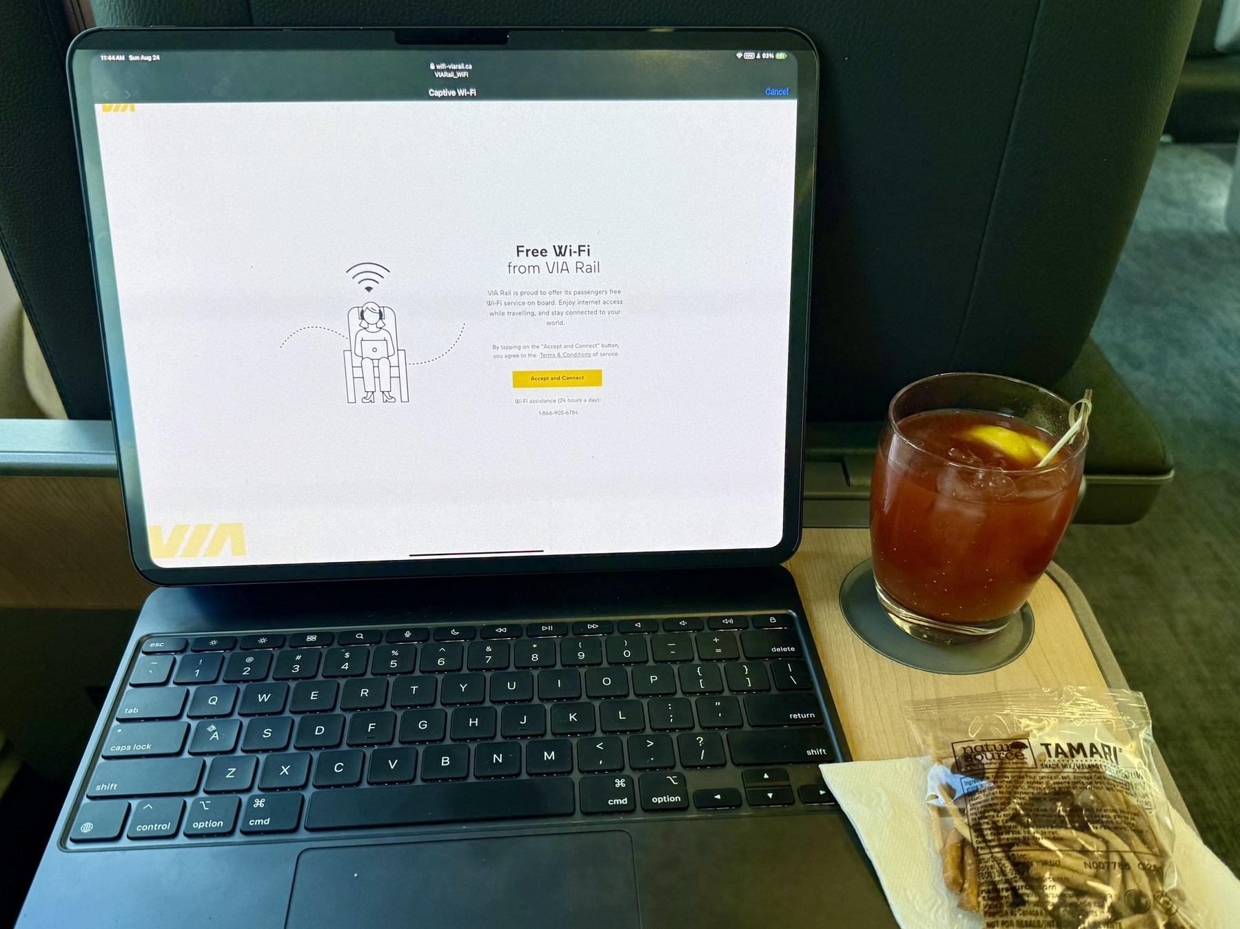 An open laptop on a train table displaying a VIA Rail free Wi-Fi login page. Next to the laptop is a glass of iced drink with a lemon slice and a wrapped package of Tamari-flavored almonds on a napkin.