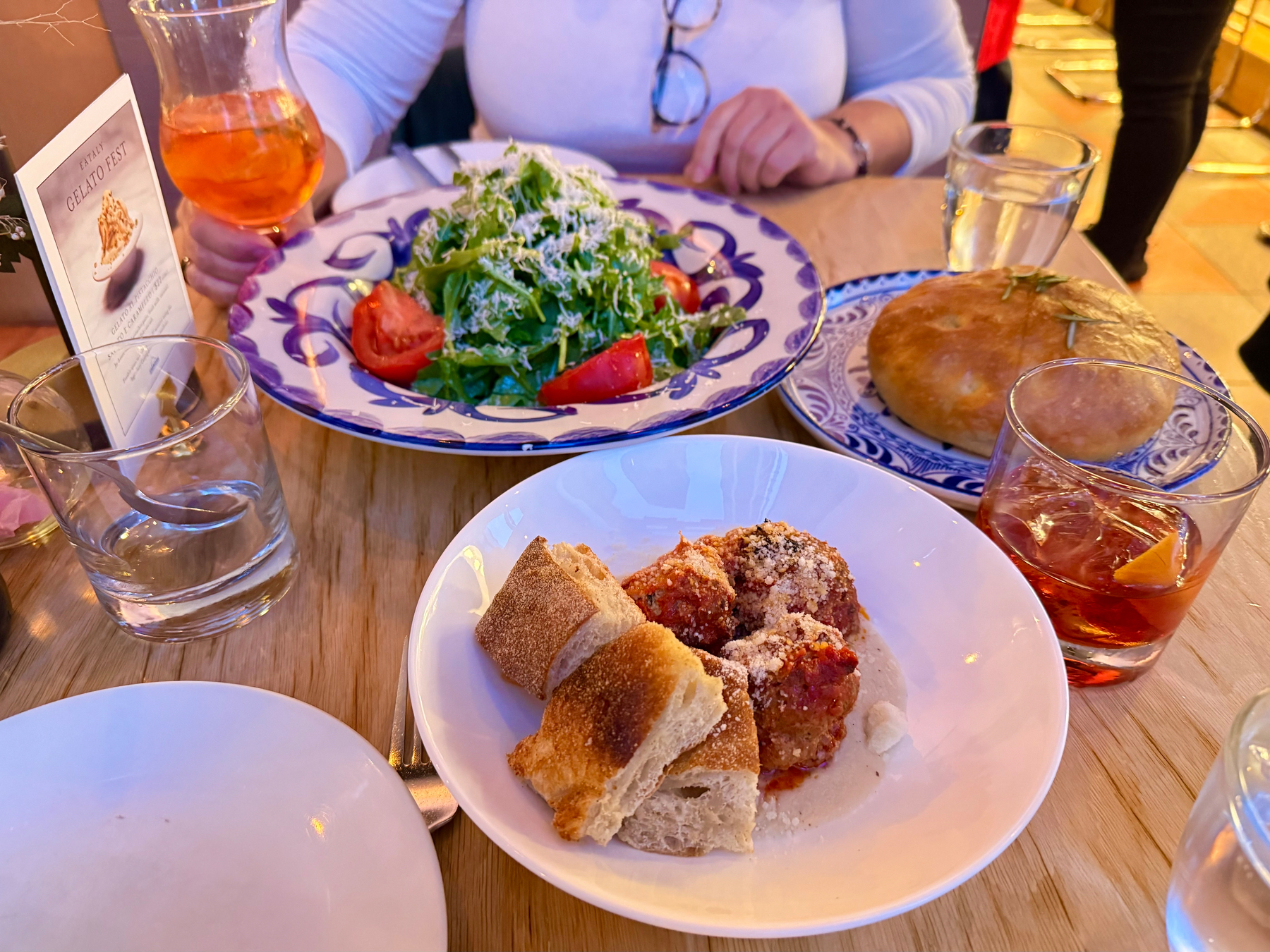 A table setting at a restaurant featuring a plate of mixed greens with tomato wedges and cheese, a round loaf of bread, and a dish of meatballs with crusty bread pieces. There is a glass of orange-hued drink, another drink with an orange slice, a glass of water, and a folded menu titled "Gelato Fest." A person is reaching for one of the drinks.
