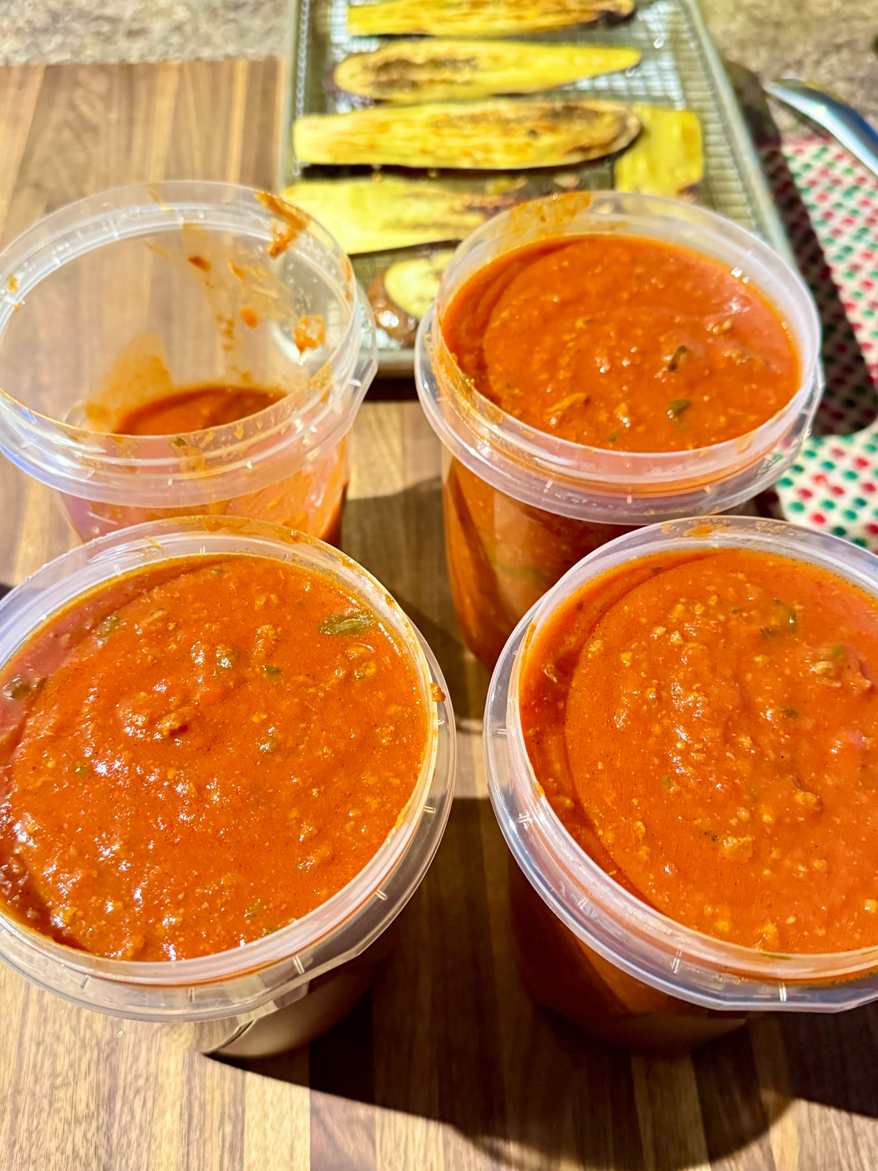 Containers filled with red pasta sauce are placed on a wooden surface. In the background, roasted eggplant slices on a baking tray are partially visible.
