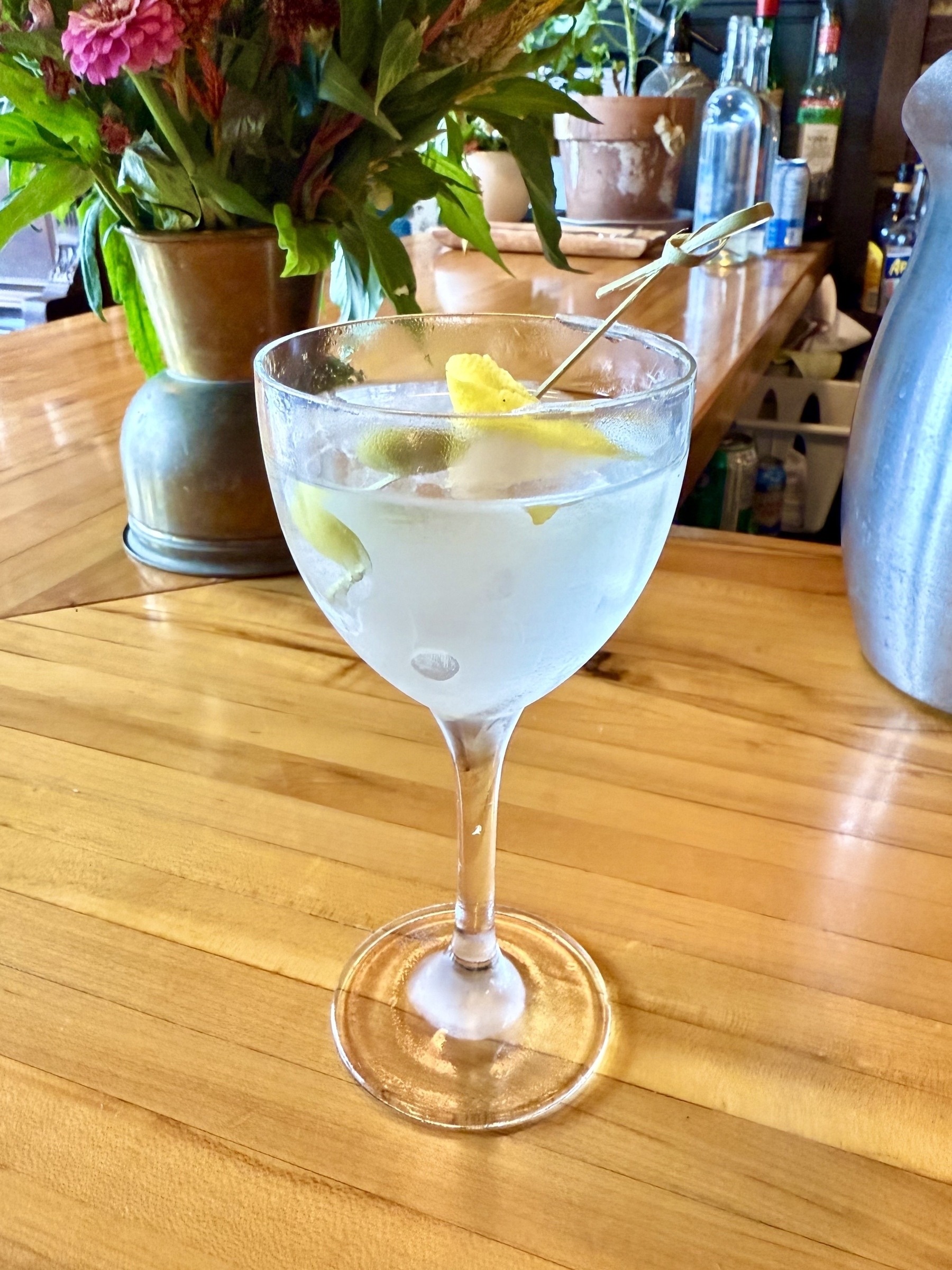 A cocktail in a stemmed glass garnished with olives and a lemon twist on a skewer, set on a wooden bar counter. In the background, there are plants in metal and terracotta pots, along with various bottles and a vase with pink flowers.
