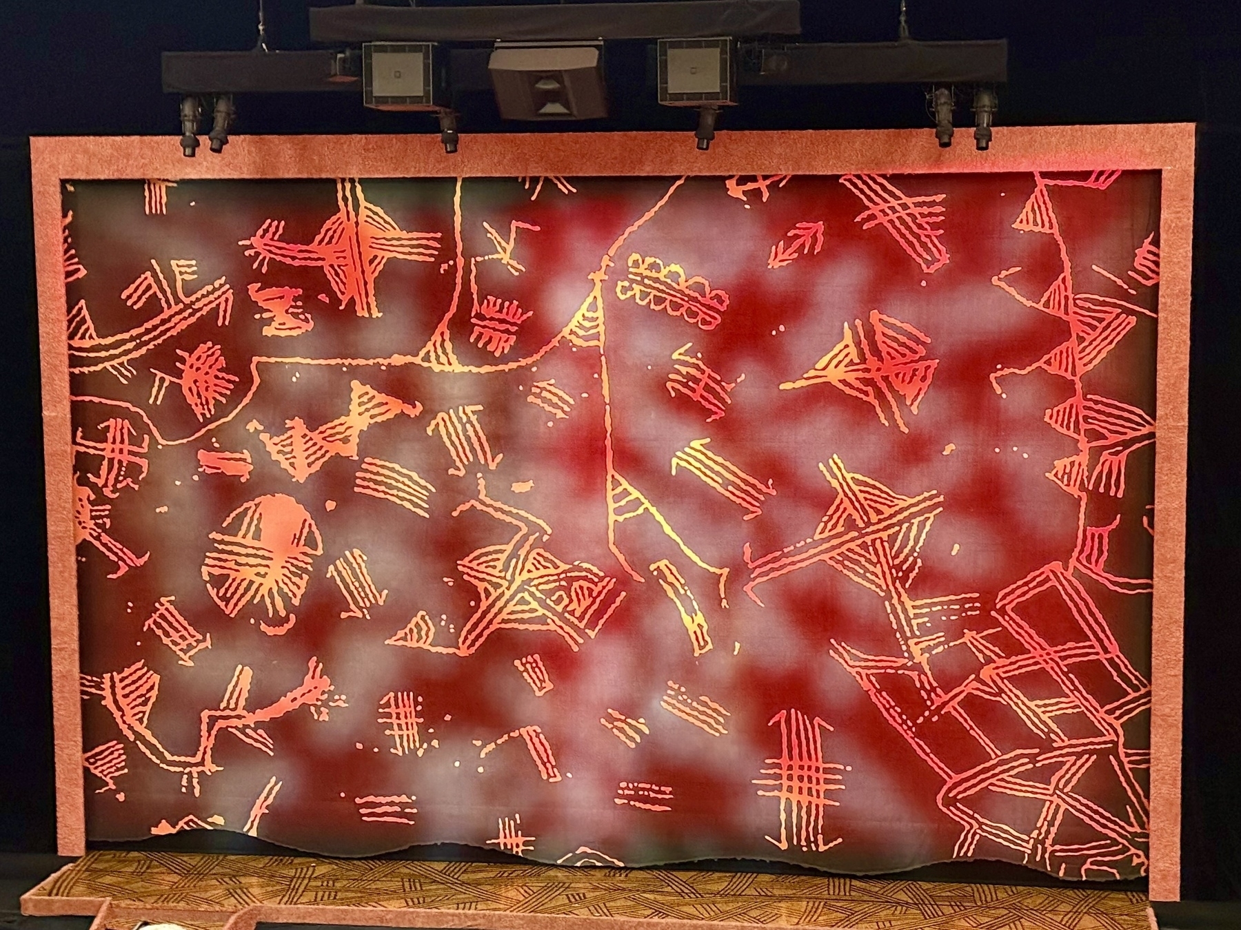 A colorful theater curtain is adorned with abstract, tribal-like patterns in shades of orange, red, and yellow on a deep red background. The designs appear geometric and organic, resembling a mix of lines, dots, and shapes. The curtain is framed by a textured border and lit from above by stage lights.