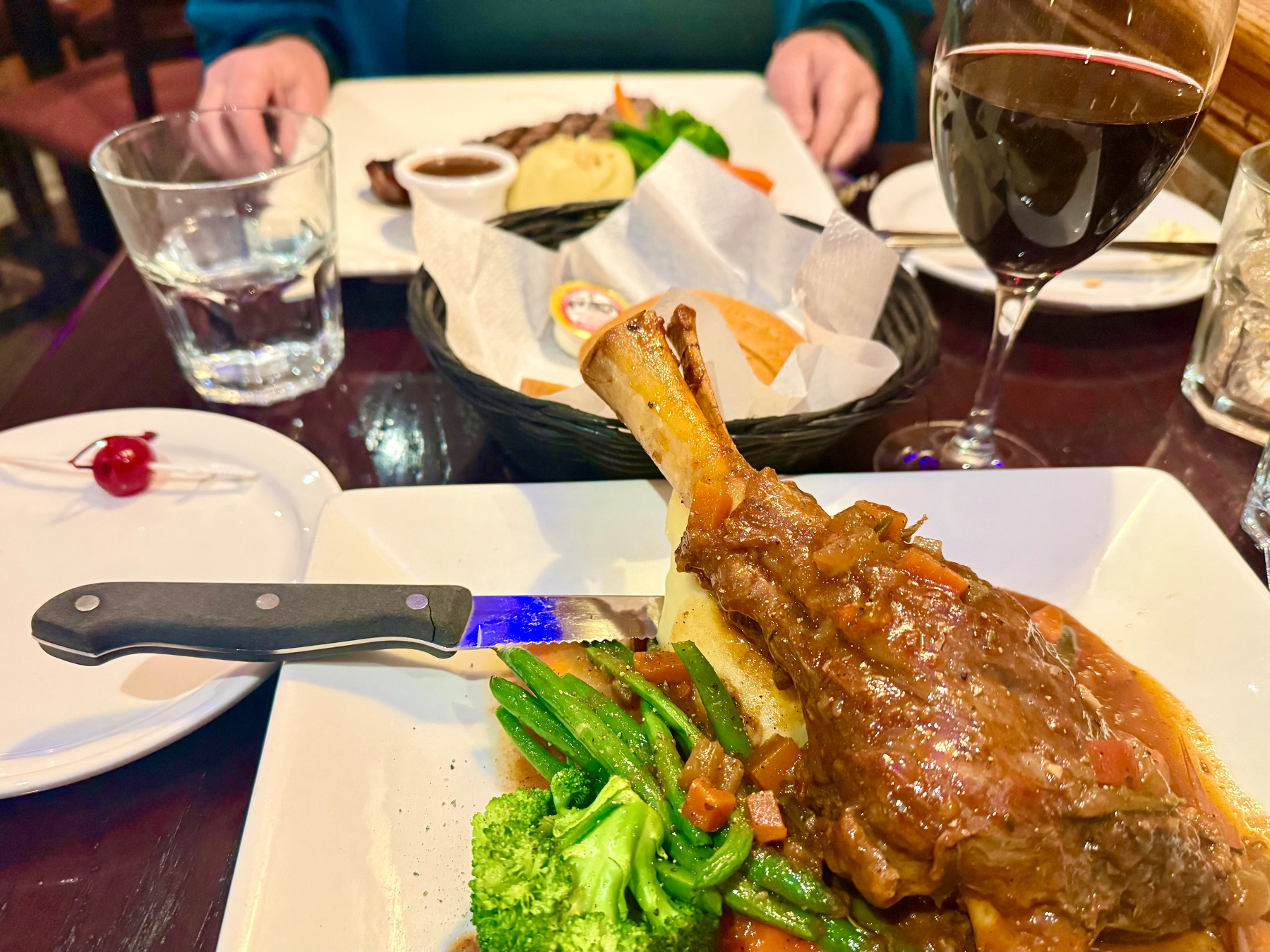 A dinner setting featuring a plate with braised lamb shank on mashed potatoes, accompanied by broccoli and green beans. A knife rests on the plate. In the background, there's a plate with steak and vegetables, a basket of bread, a glass of red wine, and a glass of water. A cherry on a toothpick is placed on a small plate in the foreground.