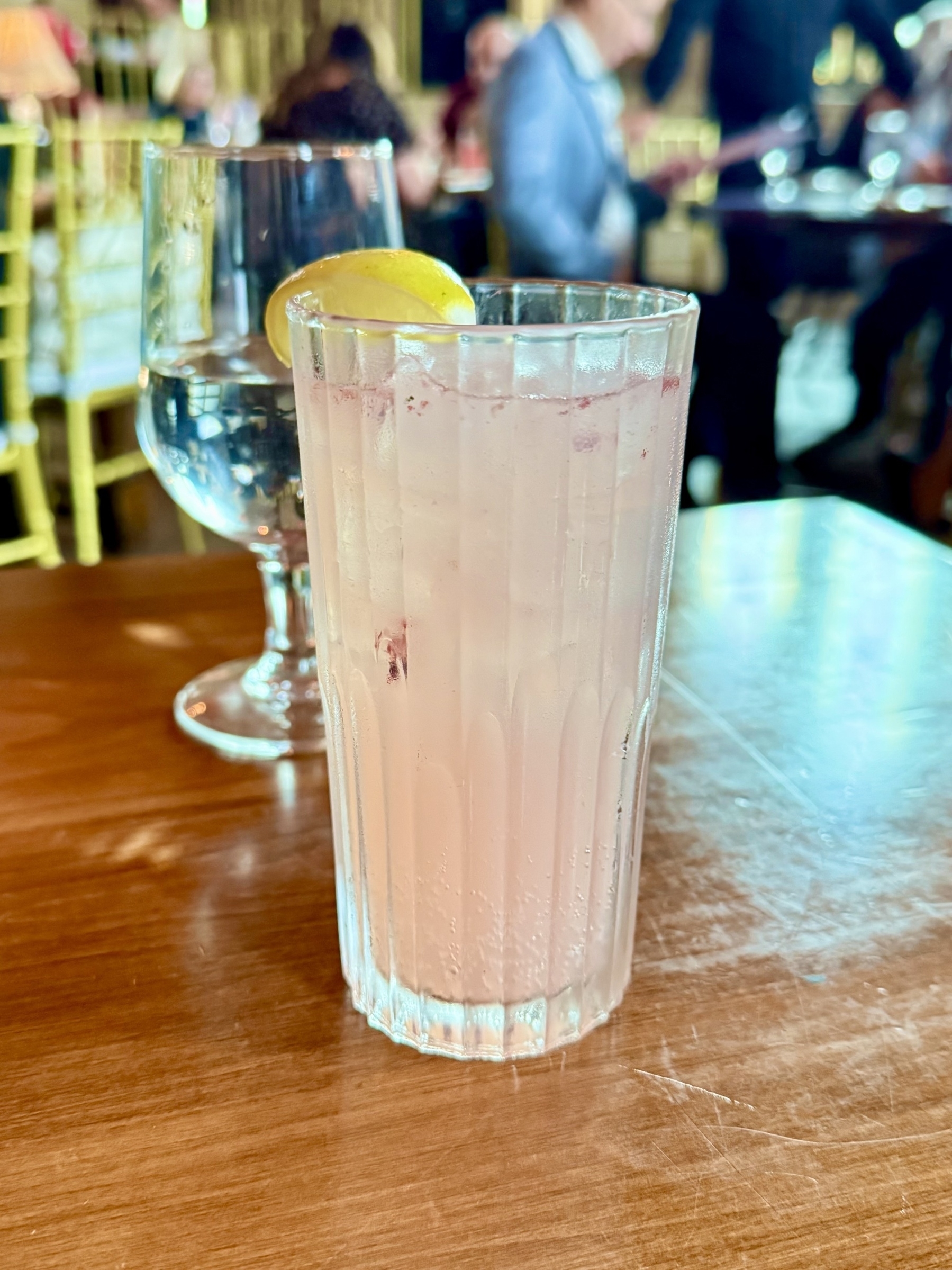 A tall glass filled with a light pink drink sits on a wooden table. The drink is garnished with a slice of lemon on the rim. In the background, there is a glass of water. The setting appears to be a restaurant with people seated and engaged in conversation, with blurred golden chairs visible.