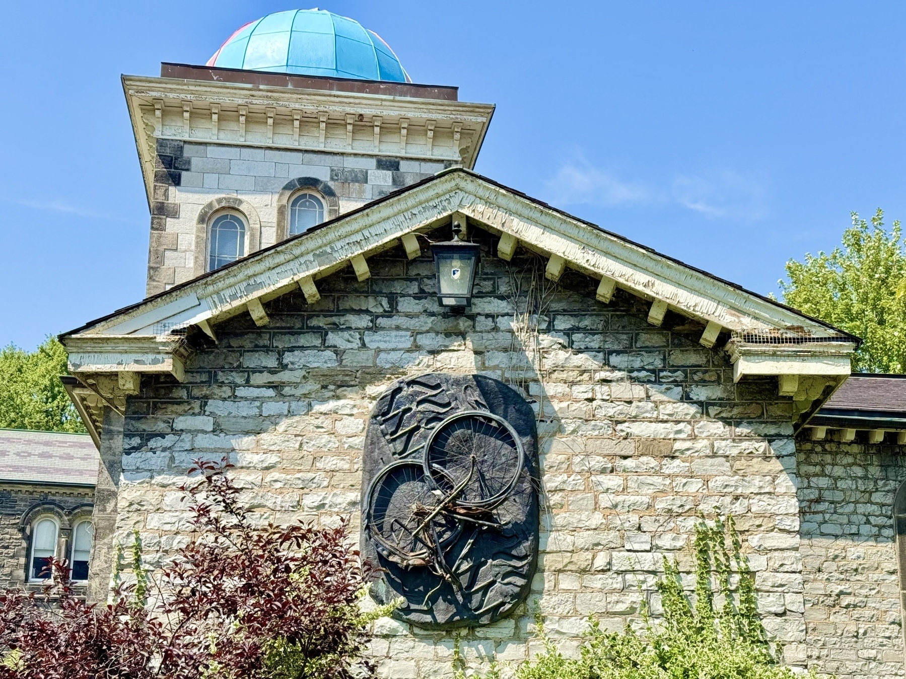 A stone building with a blue dome sits under a clear blue sky. The facade features two arched windows and a bicycle-themed sculpture mounted on it. Lush green foliage and plants are visible in the foreground.