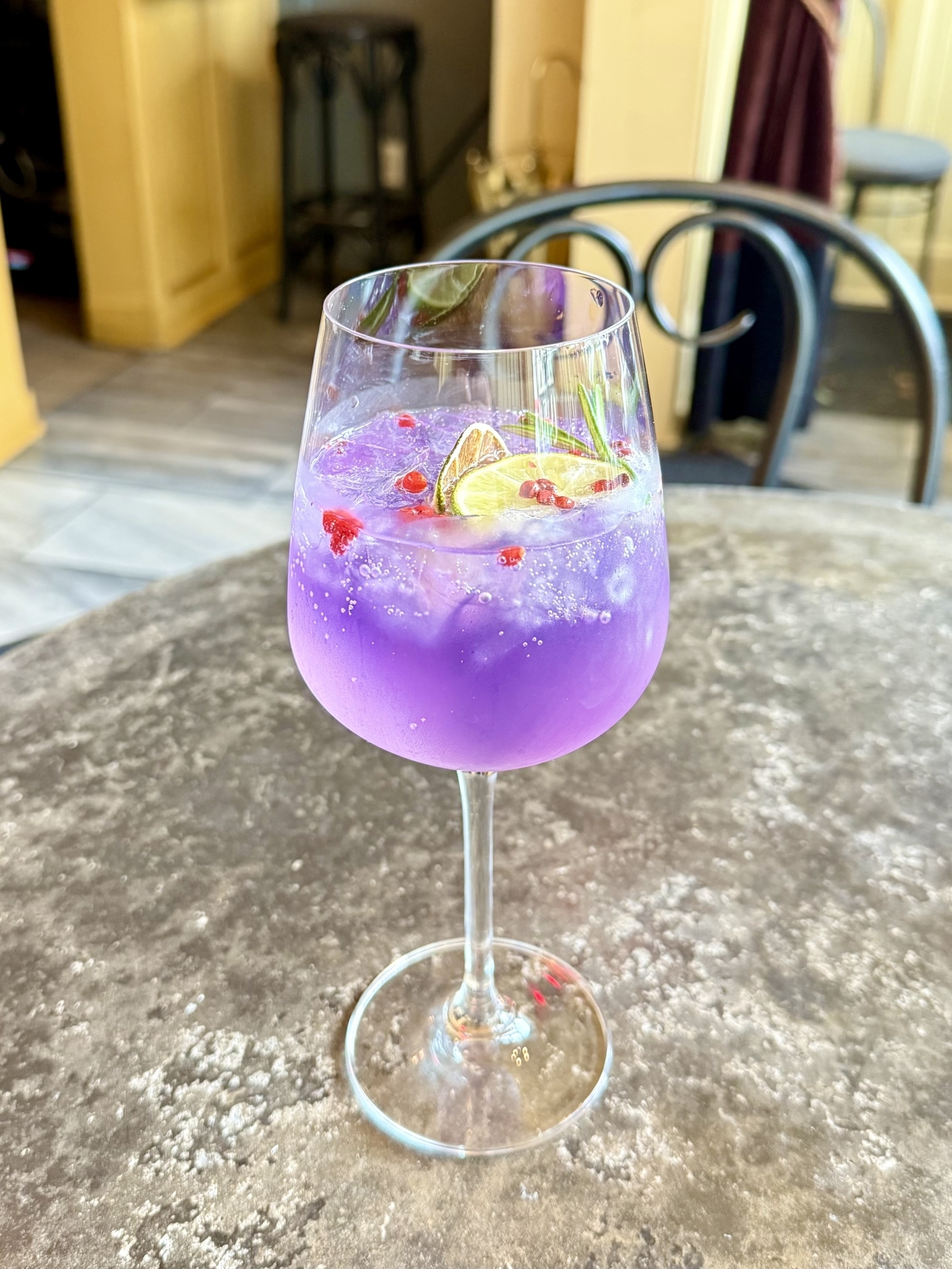 A vibrant, purple cocktail served in a wine glass, garnished with slices of lime, sprigs of rosemary, and pink peppercorns. The drink is placed on a textured, rustic table surface, with a blurred café setting in the background.