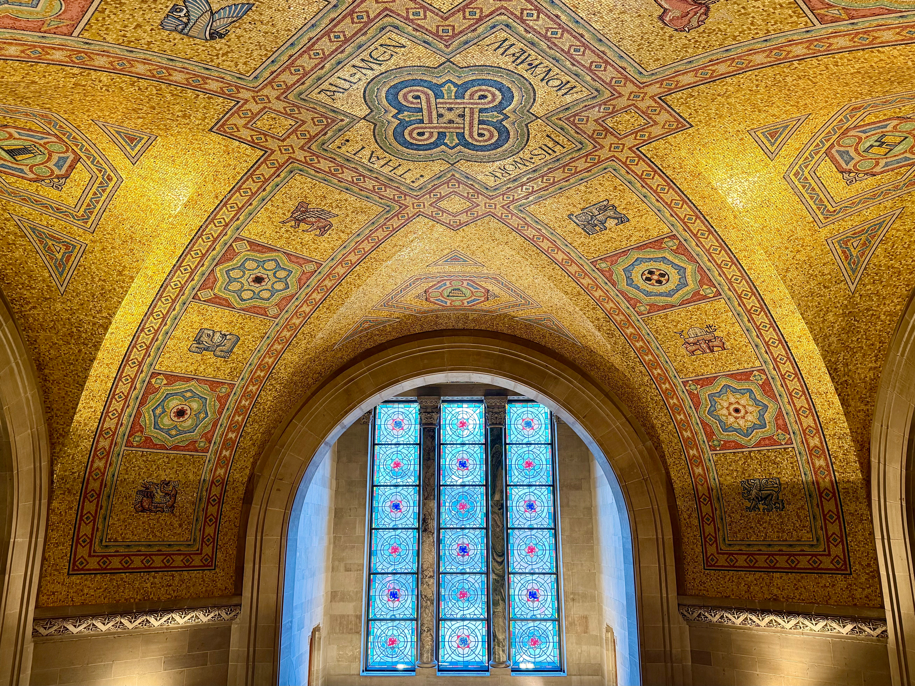 A vaulted ceiling adorned with intricate golden mosaics featuring geometric patterns and symbols, such as animals and decorative motifs. The design is highlighted with blue, red, and green accents. Below, an arched window with blue and red stained glass panels lets light into the space, emphasizing the ornate details of the architecture.