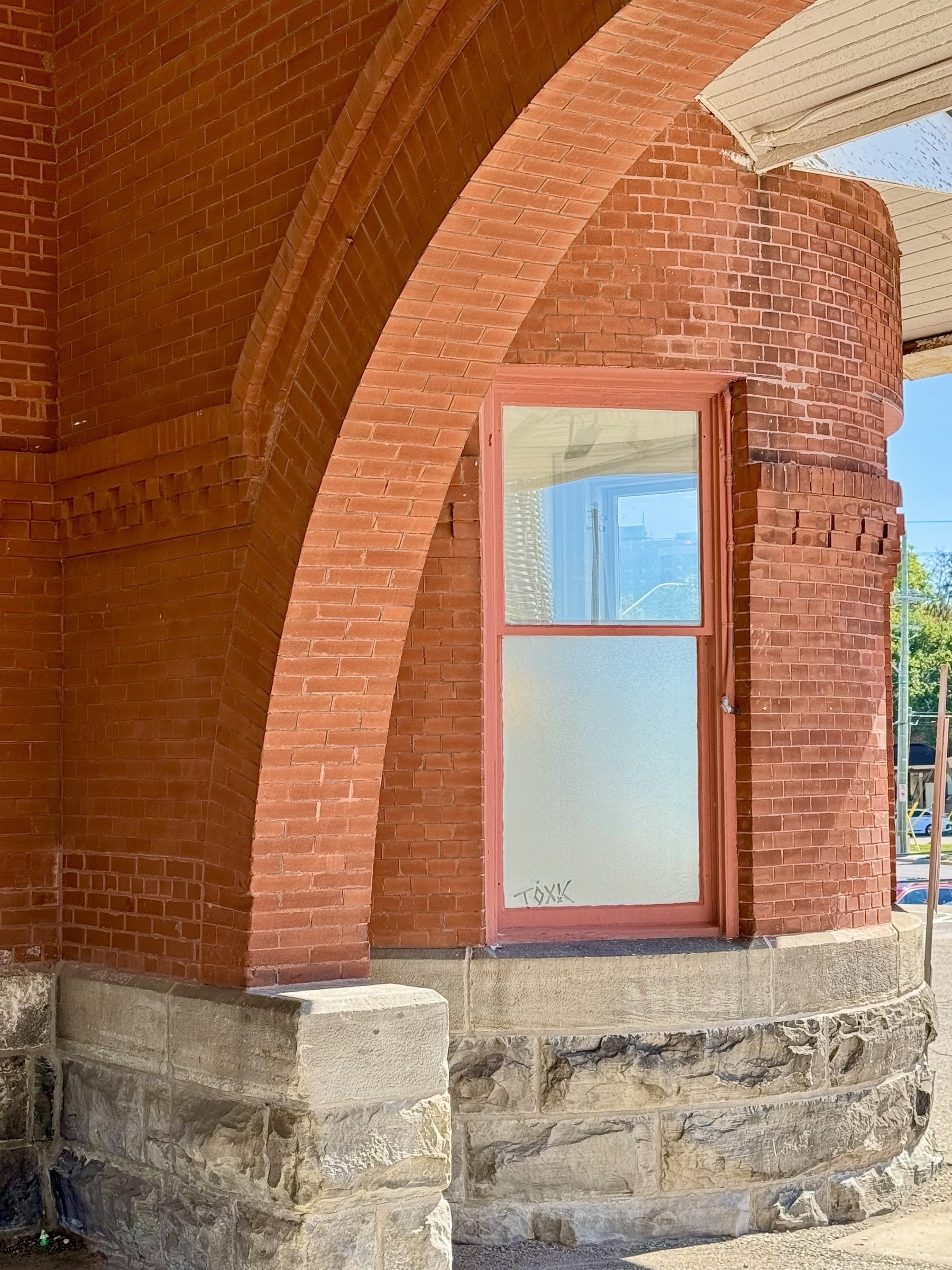 A detailed view of a red brick building showing an arched architectural design. A pink-painted window with frosted glass is set into the brick wall. The lower portion of the wall is constructed with rough grey stone blocks. Sunlight highlights the texture and color variations of the materials.