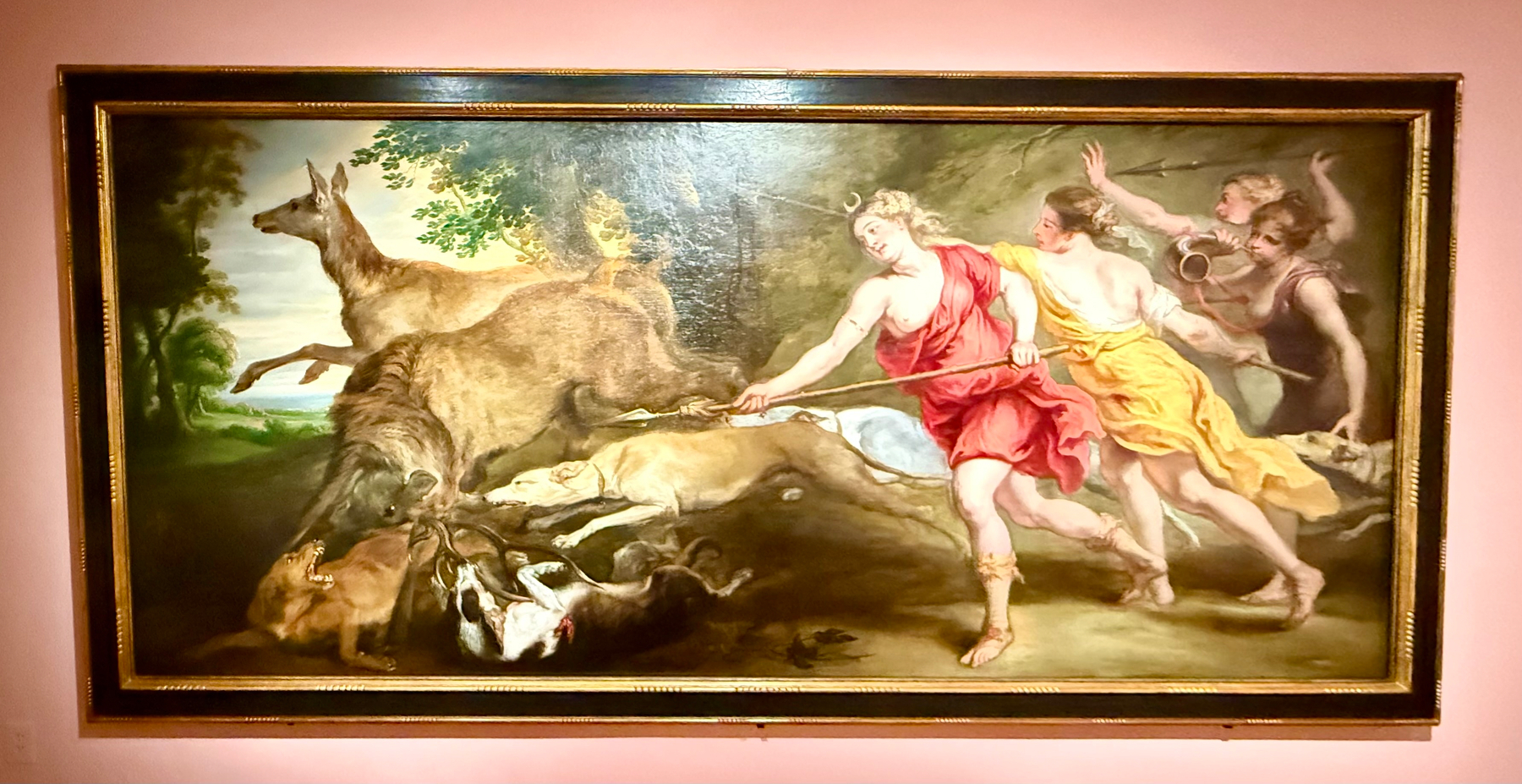 A painting depicts a dynamic hunting scene. Several figures, one wearing a red garment and another in yellow, are involved in the chase of a deer. The person in red is at the forefront, spearing an animal. Two other figures stand behind, one blowing a horn. The setting is outdoors, with trees and vegetation in the background. The painting is framed in a decorative golden frame, set against a pink wall.