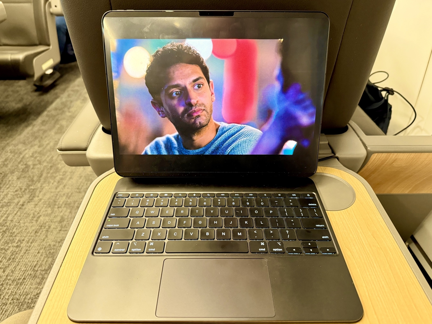 A laptop is placed on a foldable tray table in a train or airplane seat. The screen displays a scene with a person looking to the side against a colorful, blurred background. The keyboard is visible, and the setting suggests travel.