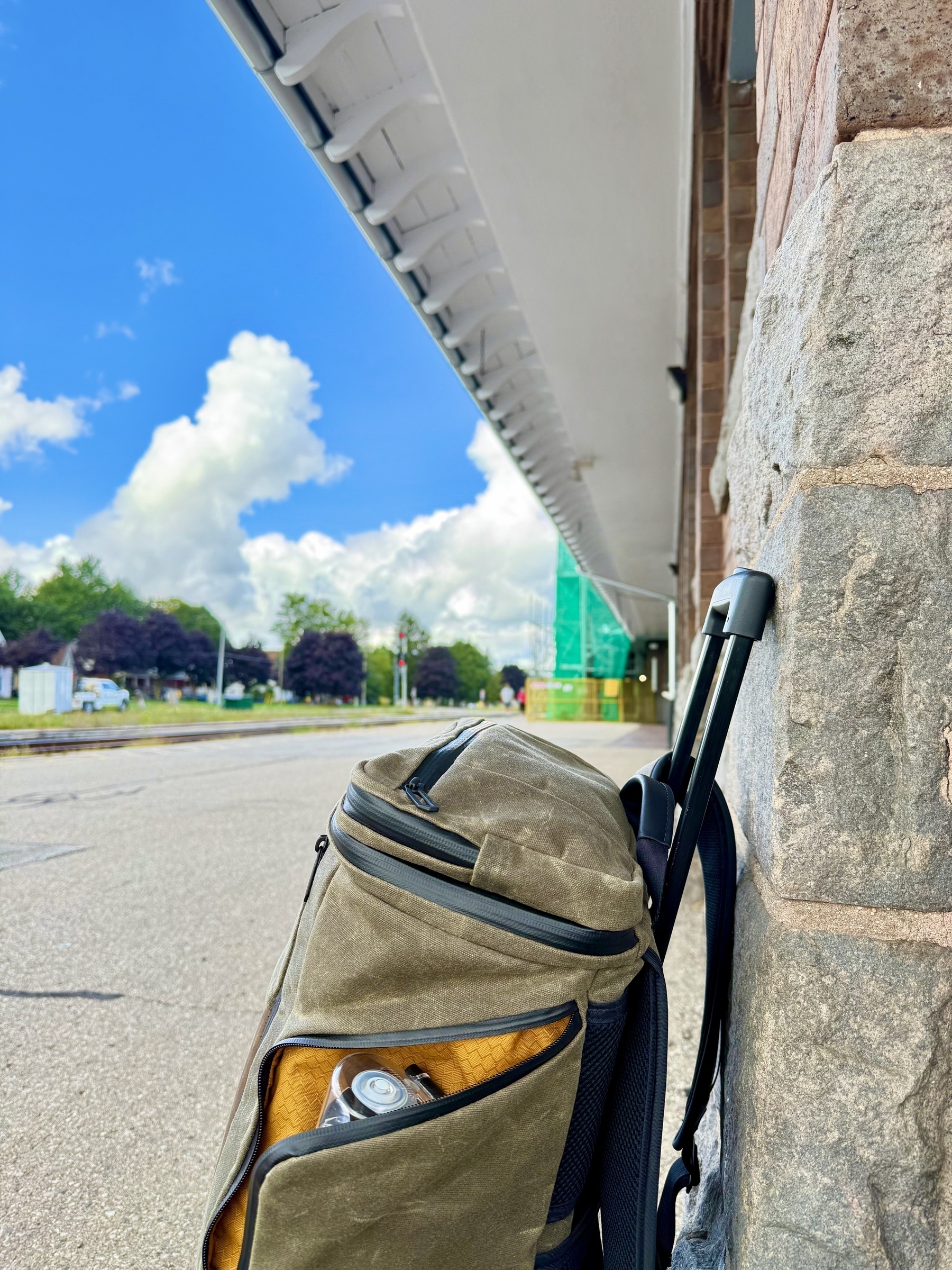 A close-up photo of a canvas backpack propped against a stone building wall. The backpack has black zippers, and a water bottle is visible in the partially open side pocket. In the background, there's an empty train platform and tracks leading into the distance. Green trees and a cloudy blue sky are visible, along with a structure covered in green netting.