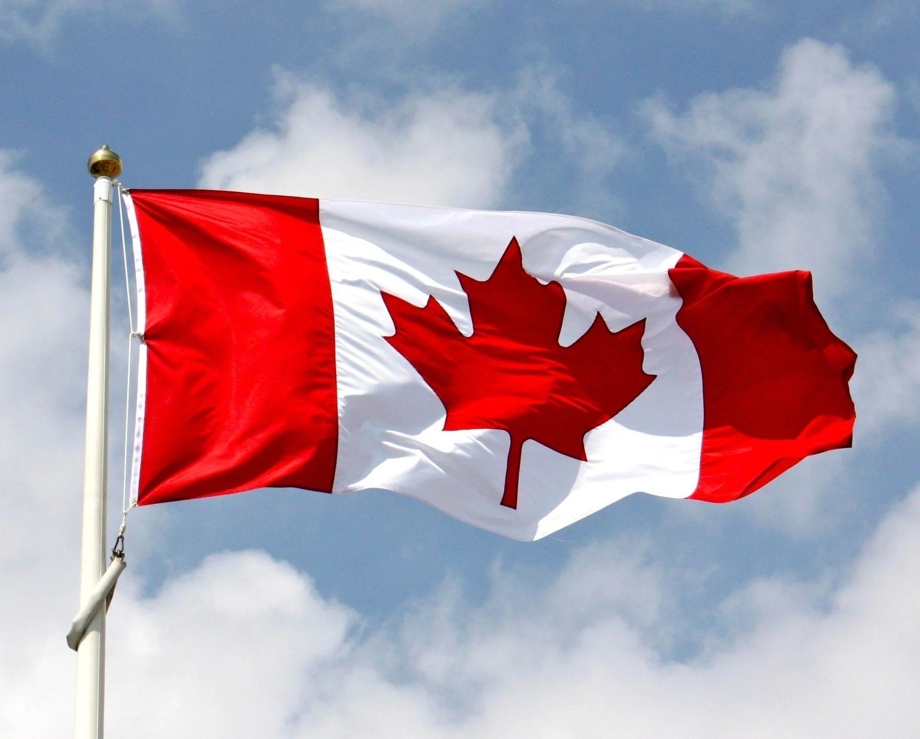 The national flag of Canada, featuring a red maple leaf centered on a white square with red vertical bands on either side, waving in the wind against a partly cloudy sky. The flag is attached to a white flagpole with a gold finial at the top.