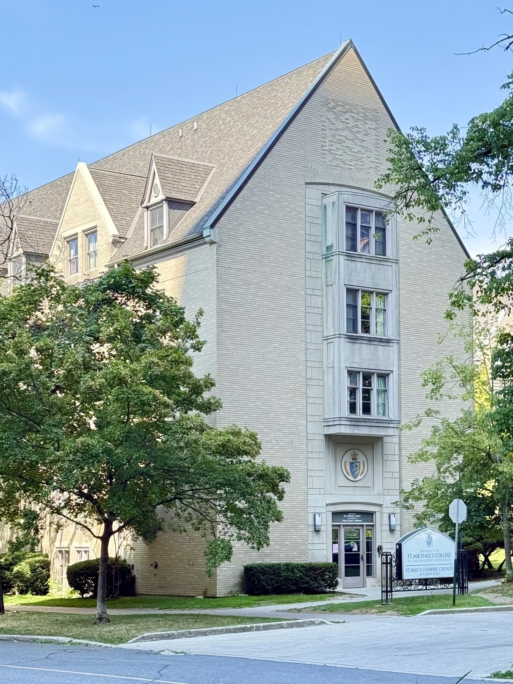 A large, multi-story brick building with a sharply peaked roof and multiple dormer windows is shown. The front of the building features a large emblem above the entrance with a coat of arms. Surrounding the building are several lush, green trees. In the foreground, there is a sign indicating "St. Michael's College," which is part of "The University of Toronto." The sky is clear and blue, indicating a sunny day.