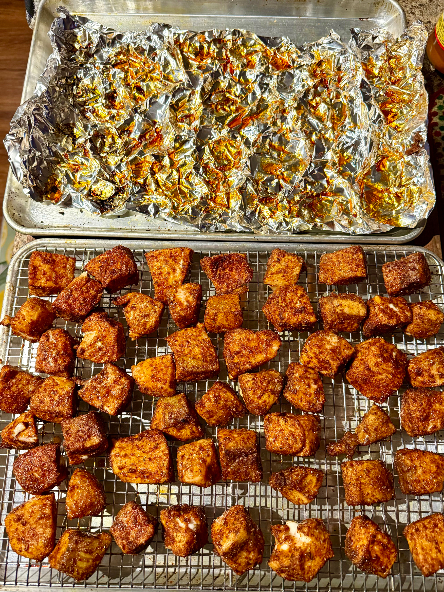 Golden-brown salmon bites are arranged on a cooling rack. Below them, a foil-covered baking tray shows remnants of roasted fat. The photo captures the crispy texture of the salmon and the remnants left from baking.