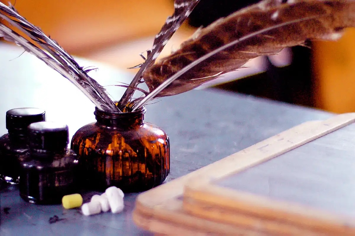 A close-up image of a vintage scene featuring several quill pens placed in a brown glass inkwell. Beside the inkwell are two small black ink bottles. In the foreground, small pieces of chalk in white and yellow are scattered on a desk. A wooden-framed slate board is visible in the bottom right corner. The background is softly blurred, adding a nostalgic feel to the image.