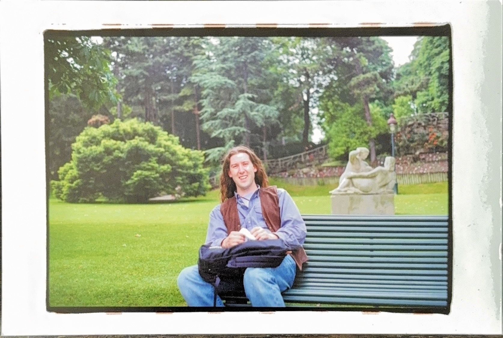 A person with long hair, wearing a blue shirt and brown vest, is sitting on a bench in a park. They are smiling and holding a bag on their lap. In the background, there's a stone sculpture and lush greenery, including trees and bushes.