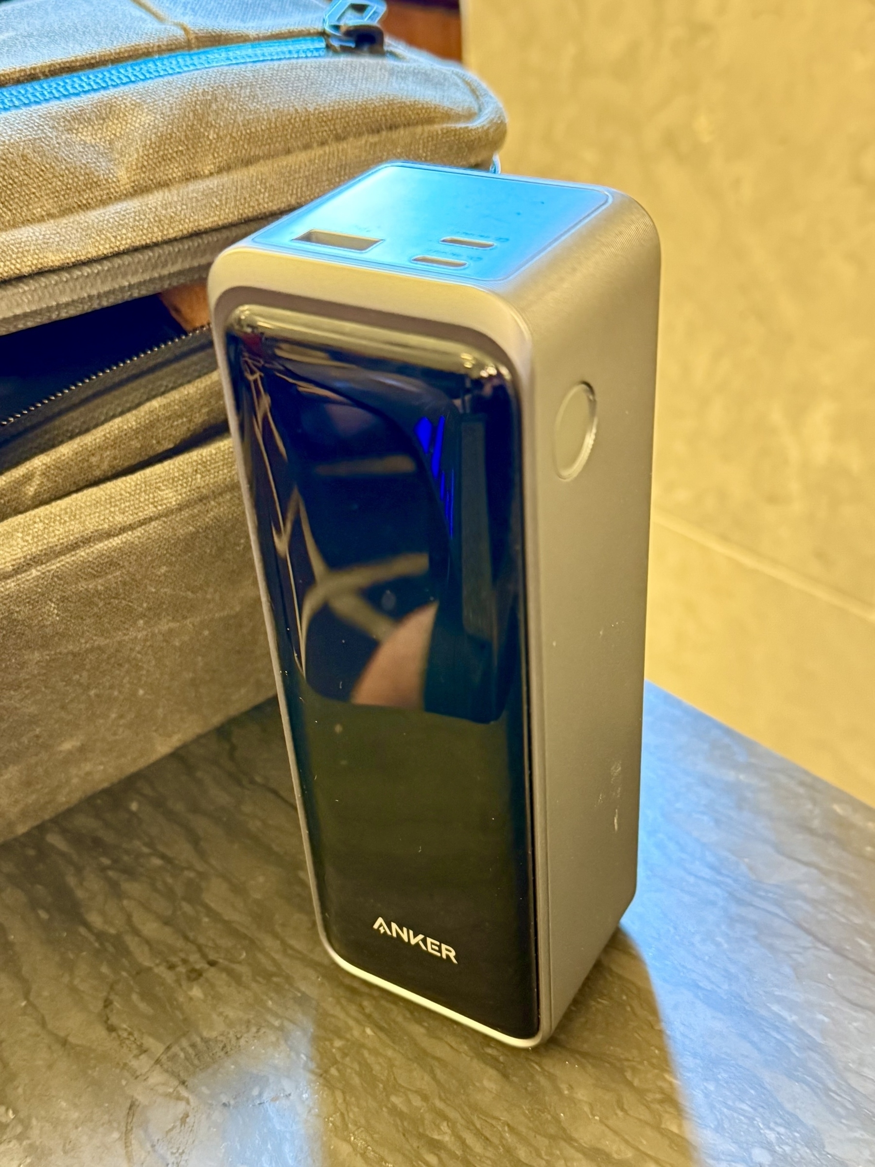 A portable Anker power bank placed on a marble surface next to a gray fabric bag with a zipper. The power bank has a sleek, metallic design with a dark glossy front, and several port openings are visible on the top.