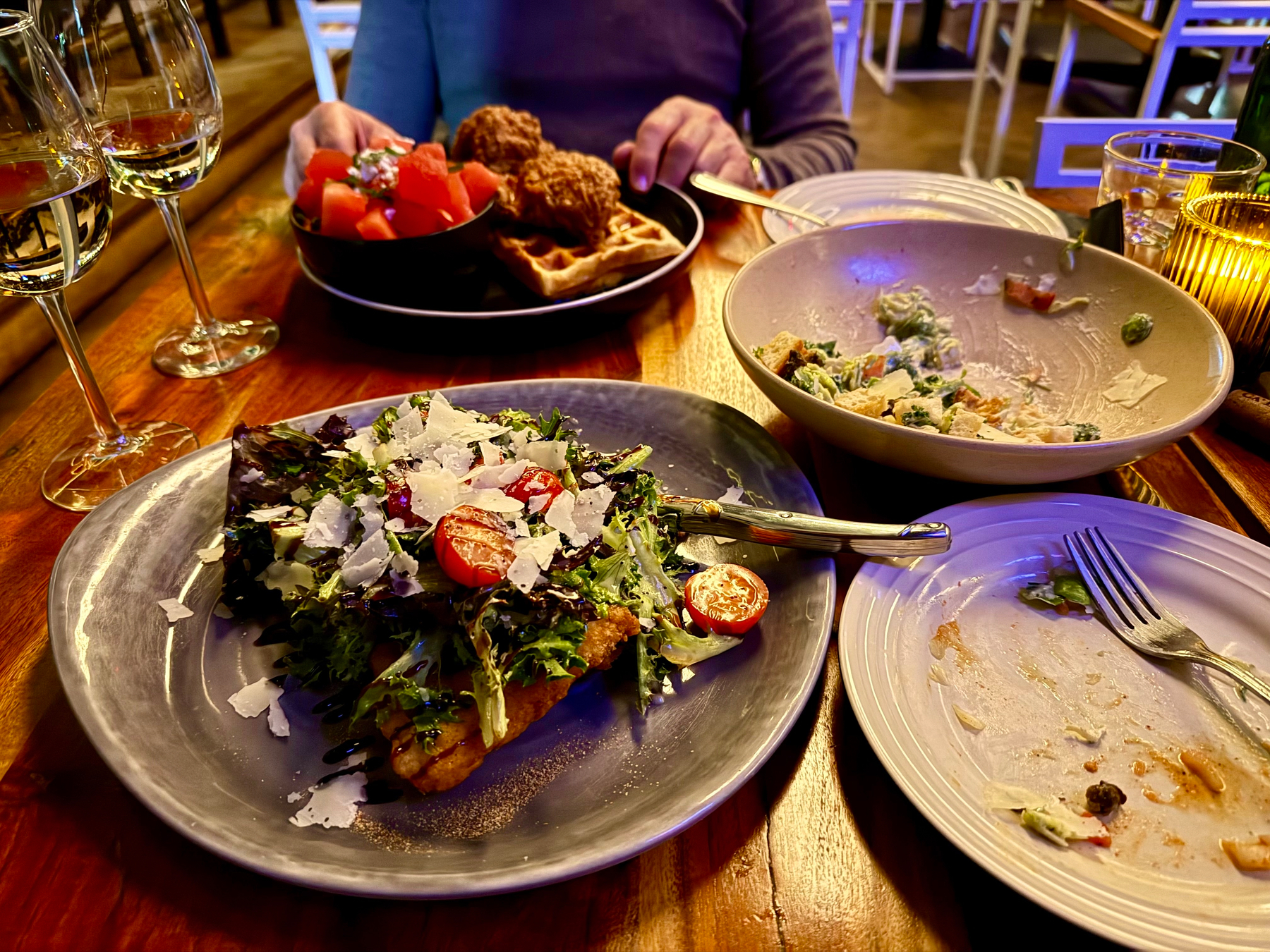 A table with a variety of dishes and drinks. In the foreground, there is a salad topped with shaved cheese and cherry tomatoes on a gray plate. To the right, a white plate and a bowl with remnants of a meal. Two glasses of white wine are in view. In the background, a person is seated with a plate containing watermelon cubes, fried chicken, and waffles. The table is wooden, and the setting is warmly lit, suggesting a cozy dining atmosphere.