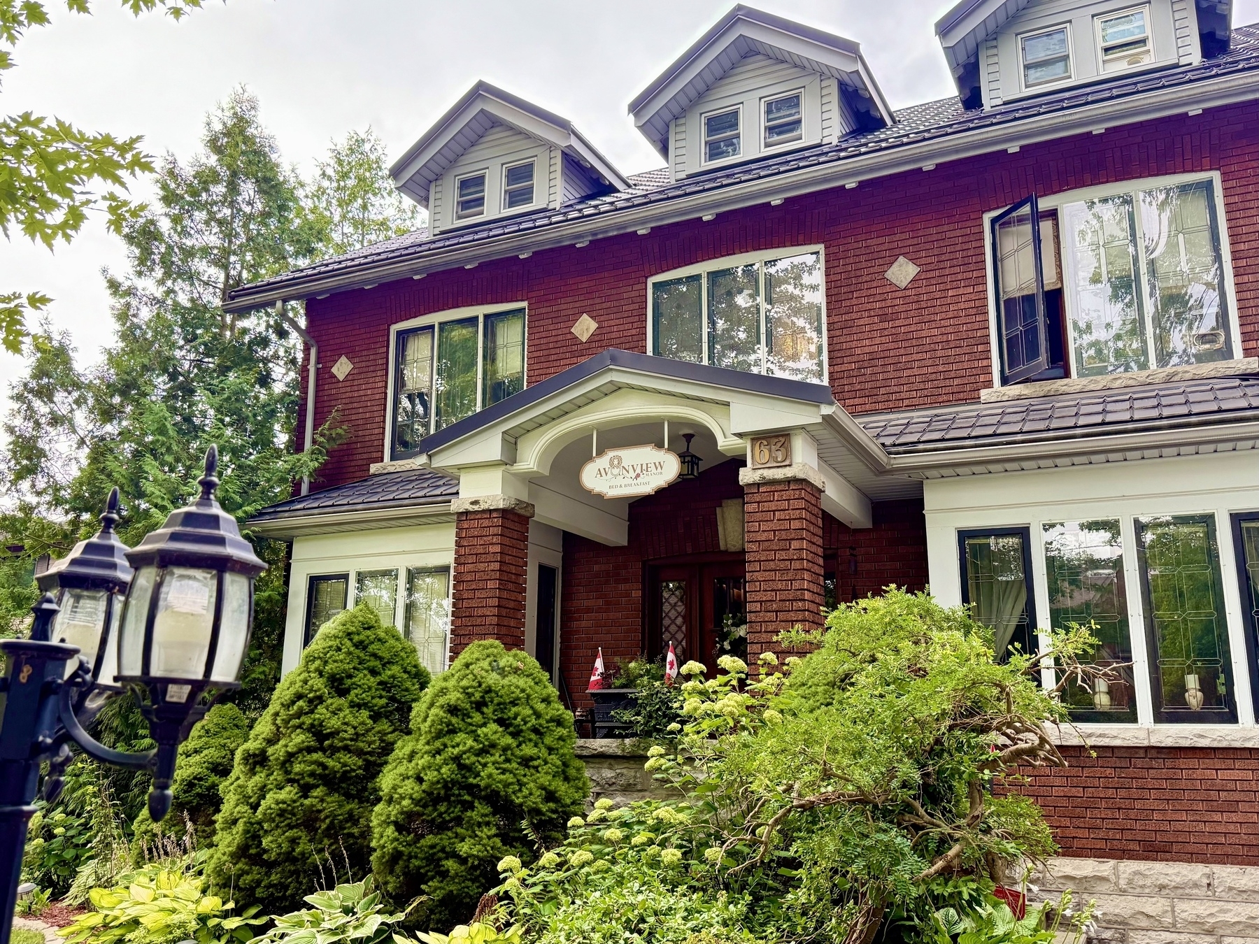 A two-story brick house with a gray roof featuring dormer windows. The entrance includes a small porch with a sign reading "Avonview" and the number 63. The house is surrounded by lush greenery including bushes and small trees. A vintage-style street lamp is visible in the foreground on the left side.