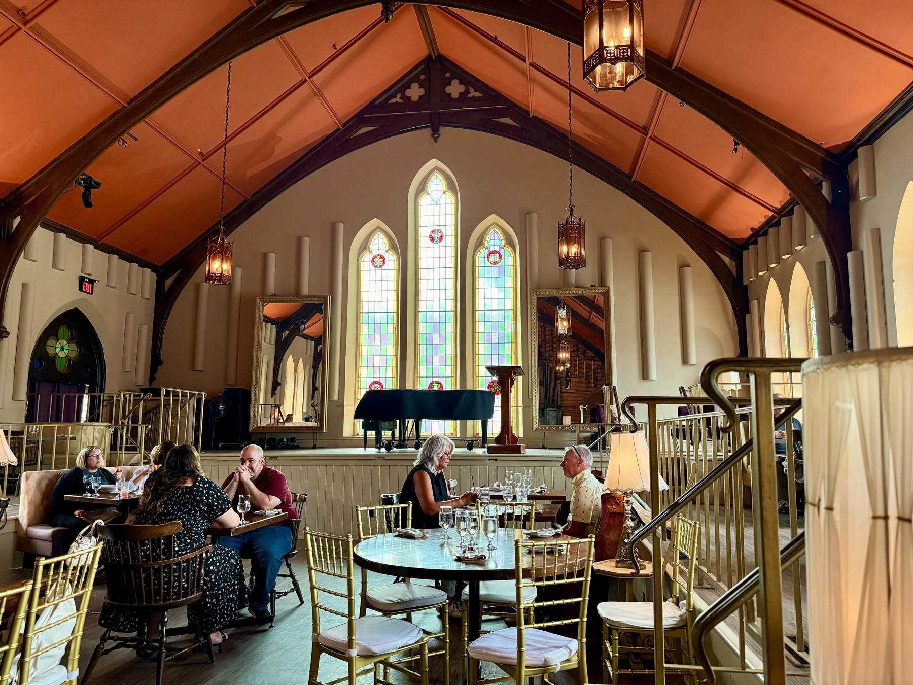 A restaurant inside a converted church with high, vaulted ceilings and large, colorful stained glass windows. Several small groups of people are seated at tables with gold chairs, enjoying their meals. A black grand piano is on a raised platform near the windows, and elegant pendant lights hang from the ceiling. The decor combines the architectural features of a church with the cozy ambience of a dining area.