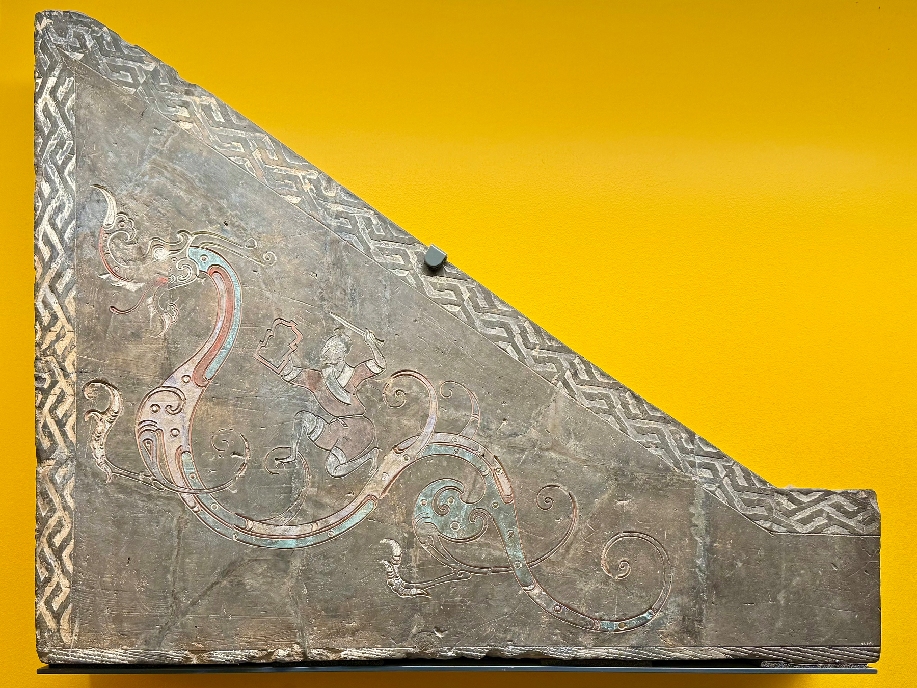 A triangular stone relief featuring an intricate design against a bright yellow background. The relief depicts a stylized dragon-like creature with curling tail and decorative patterns, alongside a human figure in the center holding a square object. The edges of the stone are adorned with a geometric border pattern.
