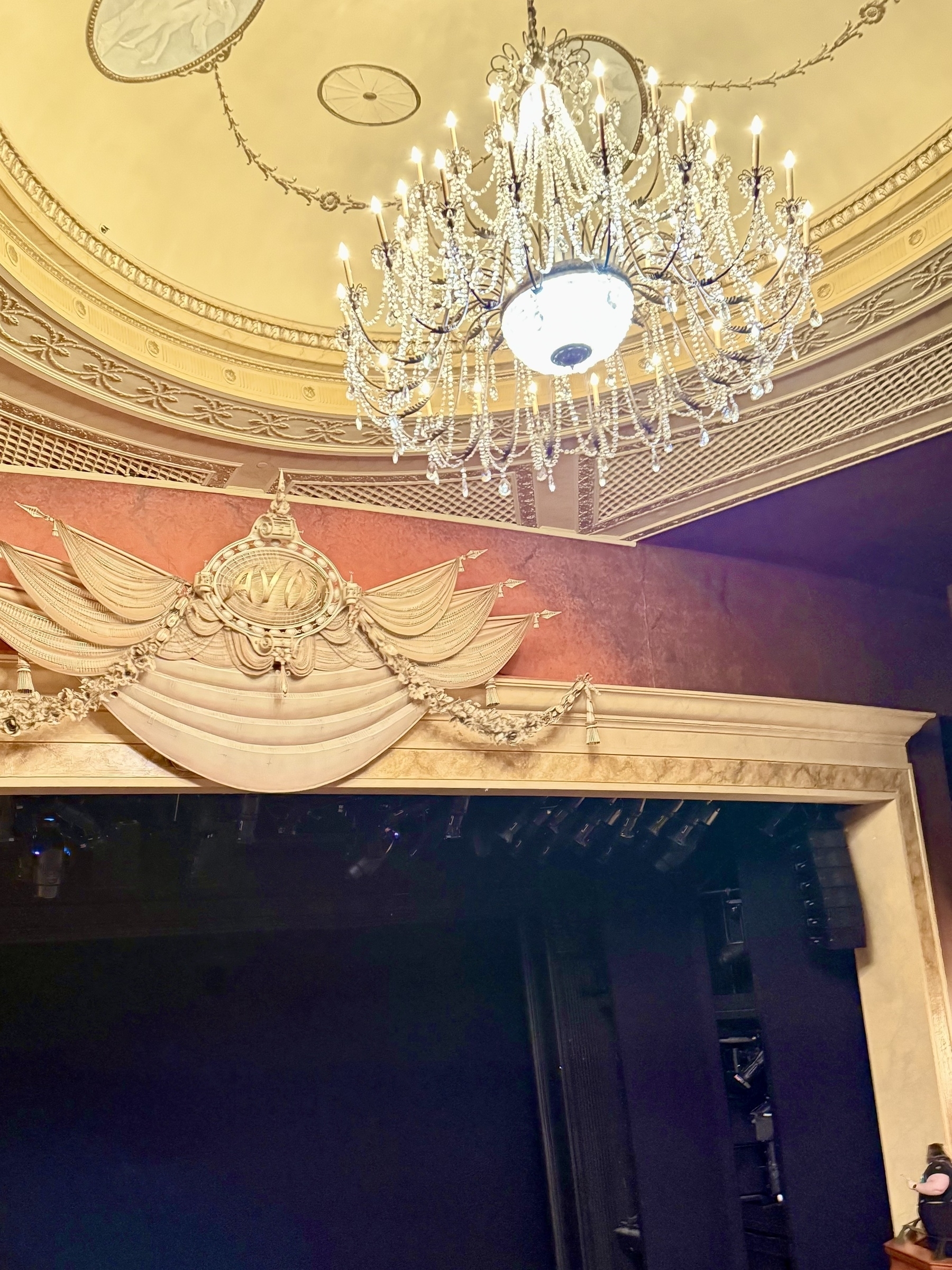 A grand theater interior featuring an ornate chandelier hanging from an intricately decorated ceiling. Below the chandelier, the stage is framed by an elegant arch with golden embellishments and drapery. The lighting creates a warm, inviting atmosphere.