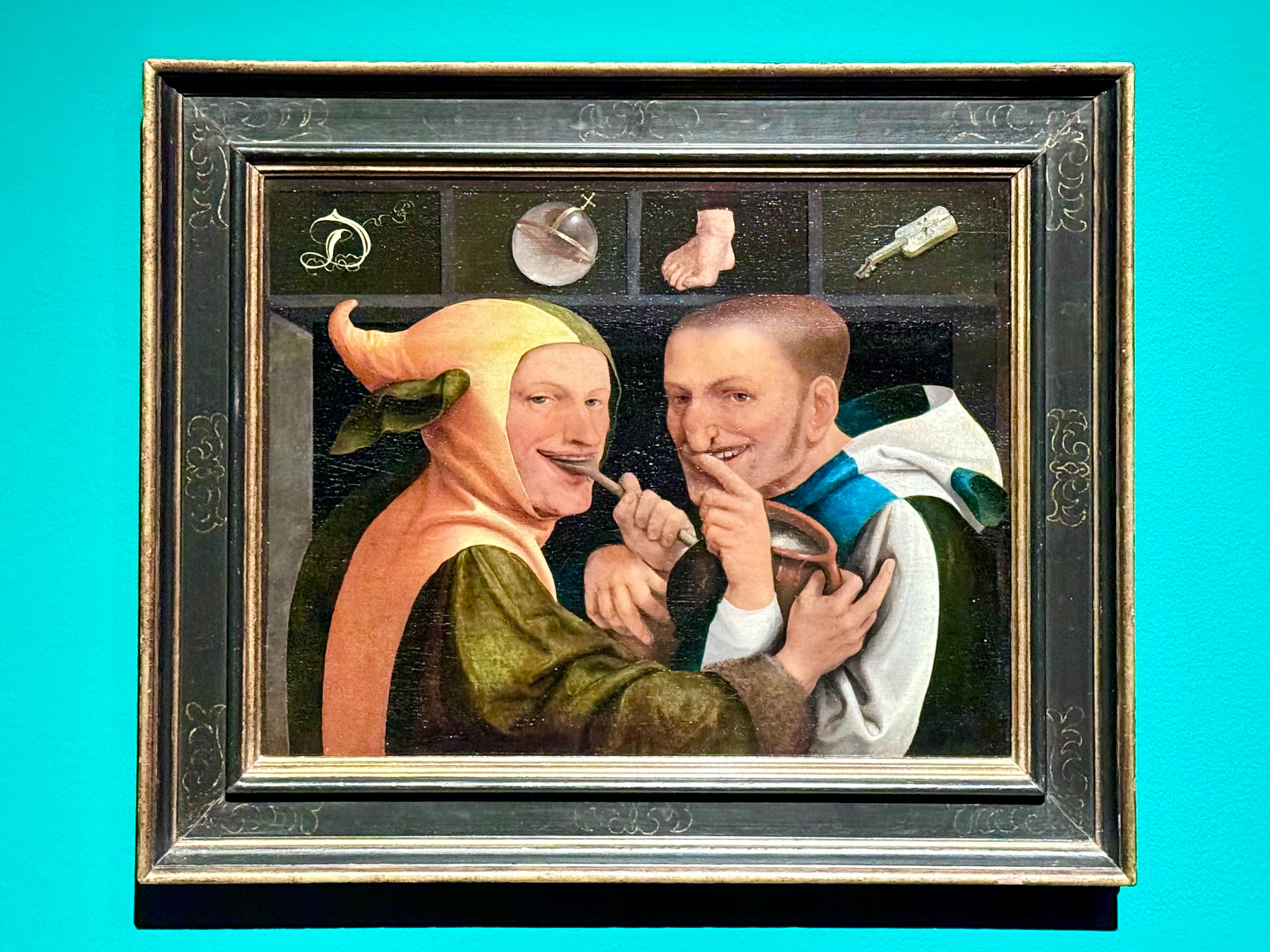 A framed painting shows two men laughing and gesturing humorously. The man on the left wears a colorful jester costume with a distinctive hat featuring a long earflap. The man on the right holds a finger to his lips, as if sharing a secret. Above them are three small symbolic elements: a dish, a foot, and a key. The background is a vibrant teal wall.