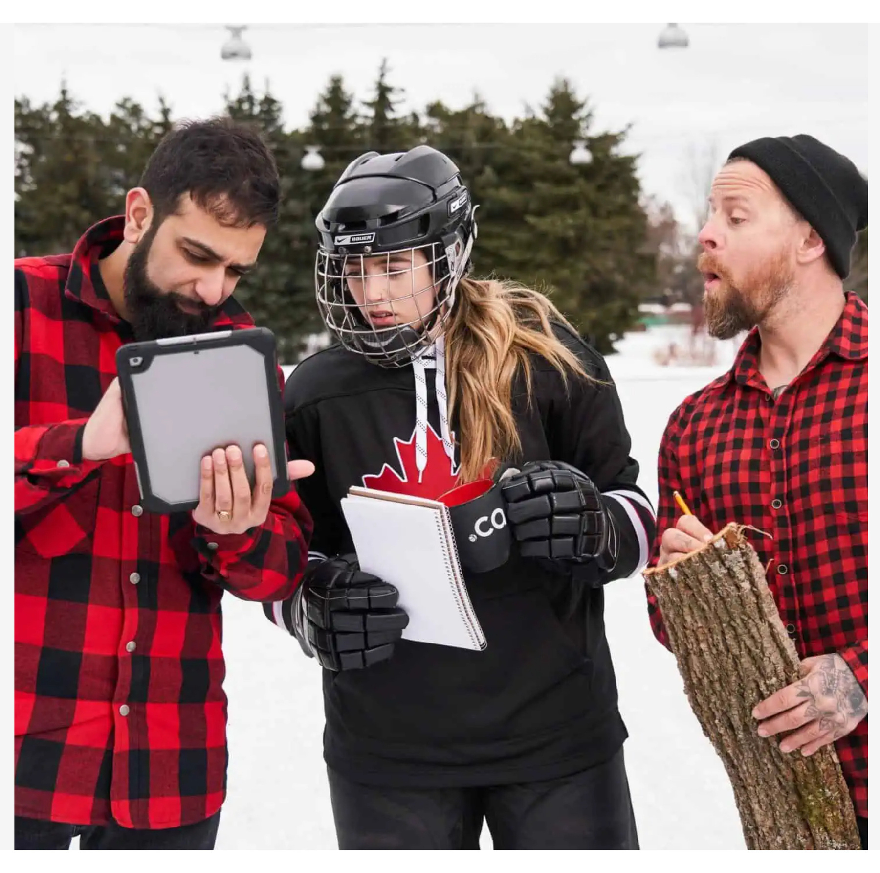 Three people are outdoors on a snowy day, seemingly engaged in a discussion. The person in the middle is wearing a black hockey jersey with a red maple leaf, black hockey gloves, and a helmet with a protective cage, holding a notebook and a black mug. To their left, a bearded man in a red and black plaid shirt is holding a tablet and pointing at the screen. To their right, another bearded man, also in a red and black plaid shirt and wearing a black beanie, holds a piece of tree bark and a pencil, appearing surprised or animated. The background features snow-covered ground, trees, and cable cars.