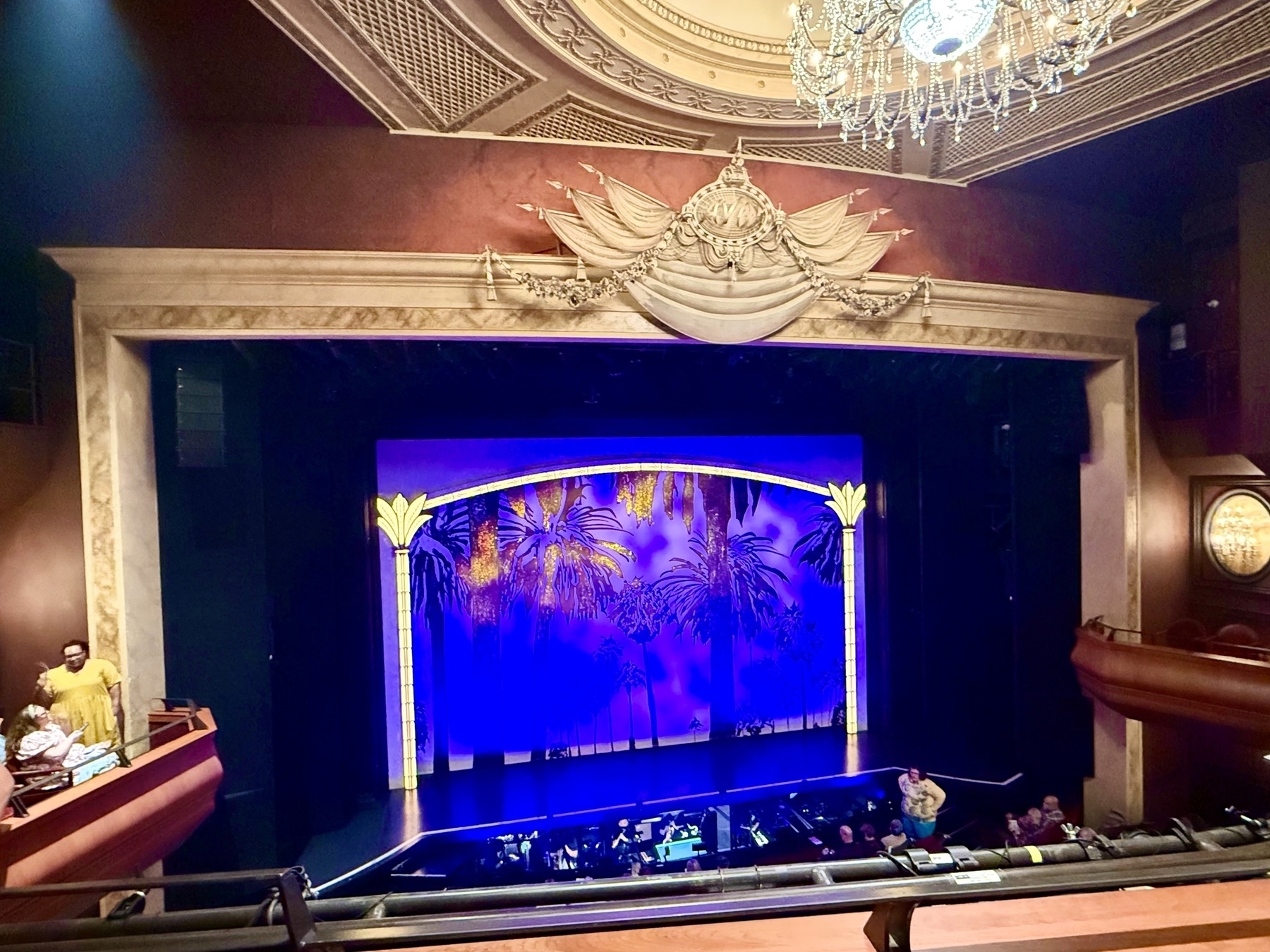 An elegant theater stage with an ornate ceiling and chandelier. The stage curtain features a vibrant blue and purple tropical scene framed by decorative columns. People are visible in the balcony and front rows, waiting for a performance to begin. The overall atmosphere is grand and anticipatory.
