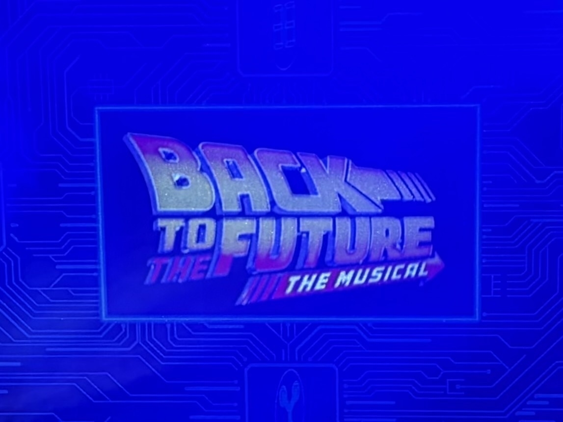 A stylized poster for "Back to the Future: The Musical" is displayed. The background features a digital circuit design in shades of blue, highlighting the words "Back to the Future" prominently in metallic letters, with "The Musical" written below in white text over a red and blue striped arrow.