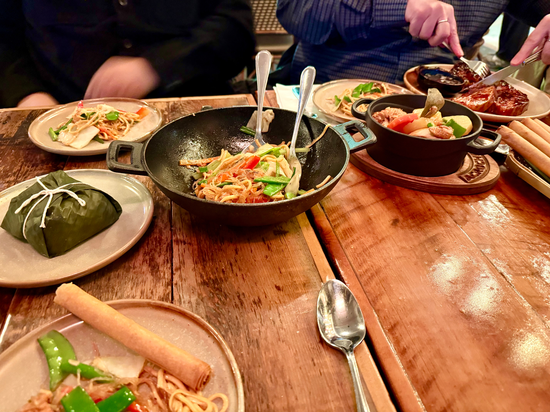 A table set with a variety of Asian-inspired dishes. In the center is a bowl with stir-fried noodles featuring vegetables like bell peppers and snap peas. Surrounding it are plates with items, including a wrapped leaf package, a spring roll, and more noodles. To the right, a small pot holds a vegetable and meat dish. Two people are partially visible, one preparing to eat. The scene is set on a wooden table.