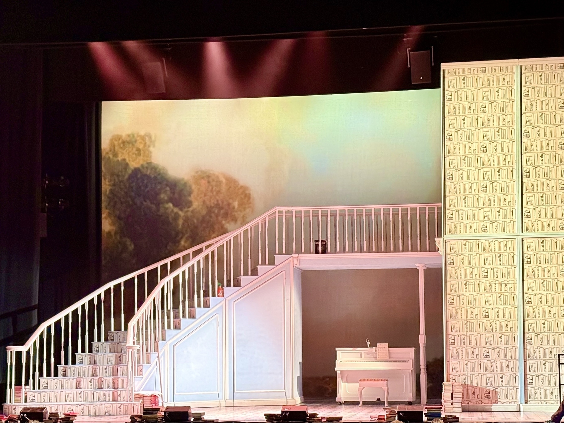 A theater set featuring a large staircase with white railings leading up from left to right. The steps are patterned with book designs. Below the staircase is a recessed area with a white piano and sheet music on top. The background displays a softly lit mural of trees. To the right of the set, there is a tall wall covered in a book pattern. The stage lighting casts a warm glow across the scene.