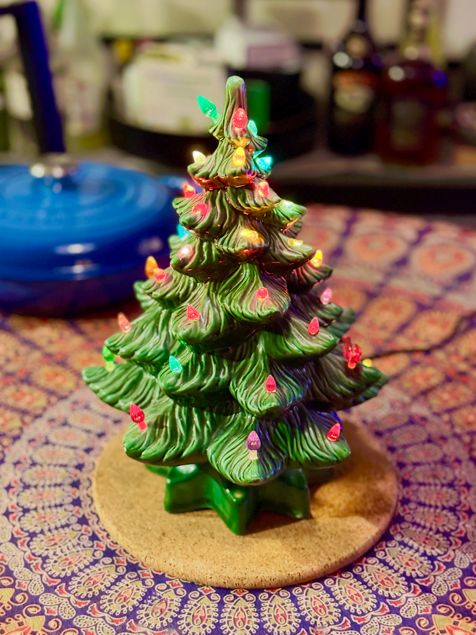 A ceramic Christmas tree with colorful lights is displayed on a decorative circular mat with a detailed pattern. The tree is surrounded by various household items that are out of focus in the background, including a blue kitchen item.