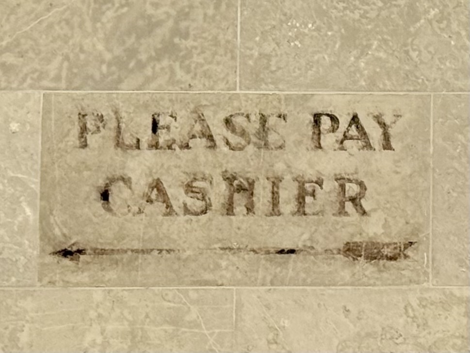 Old and faded text on a stone wall reads "PLEASE PAY CASHIER," with decorative arrows pointing to the left.