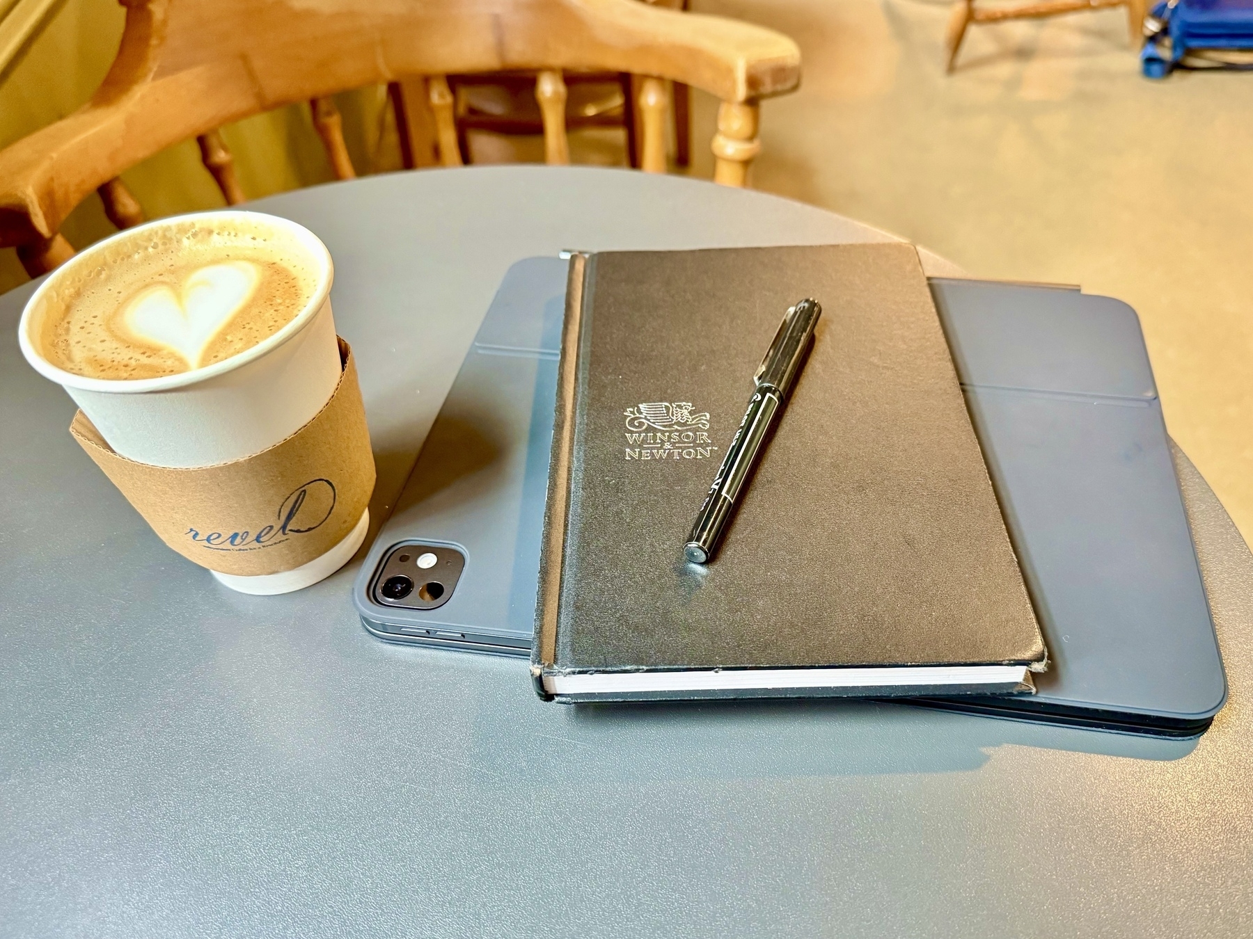 A small round table with a paper cup of coffee topped with latte art in the shape of a heart, a black notebook labeled "Winsor & Newton" with a pen on top, and a partially visible tablet device underneath. A wooden chair is in the background.