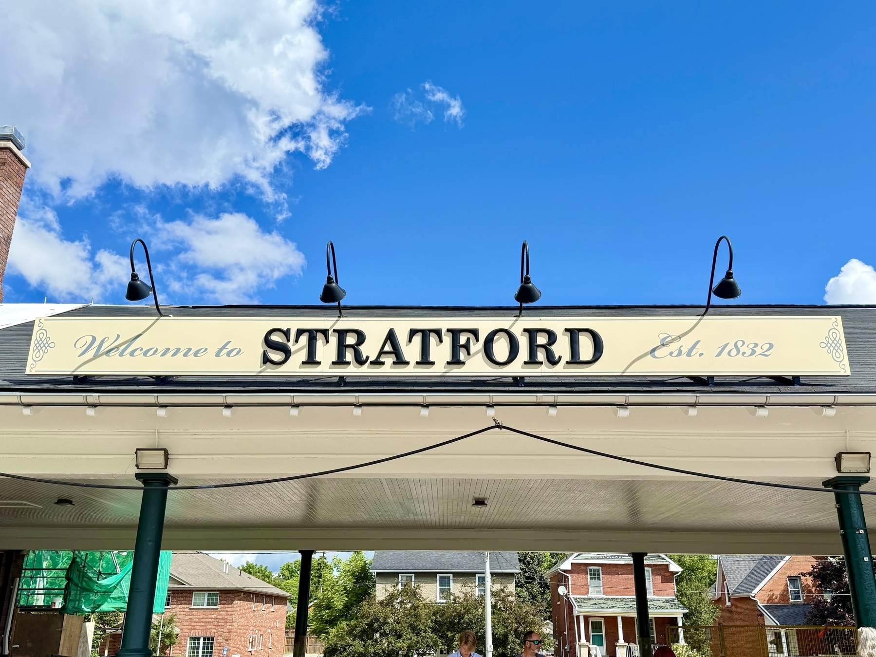 A sign with the words "Welcome to STRATFORD Est. 1832" mounted on a structure with multiple black lamps above it. Below, some brick buildings are visible in the background. The sky is bright blue with a few clouds.