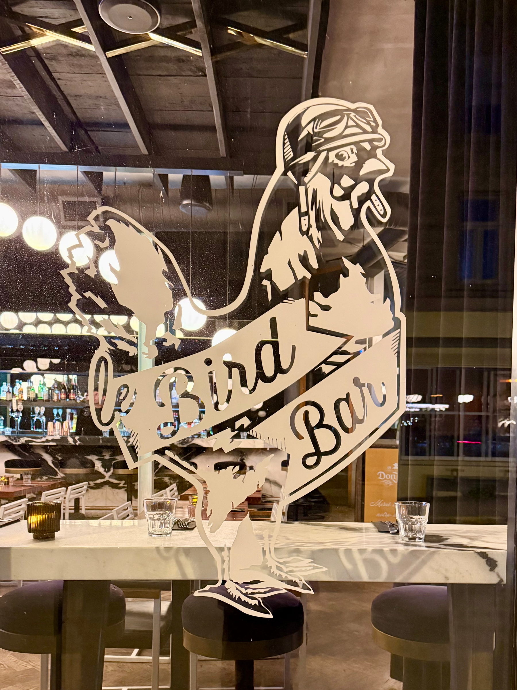 A window sign featuring an illustration of a rooster wearing aviator goggles and a scarf, with the text "Le Bird Bar" diagonally across its body. The interior of a bar is visible through the glass, with bar stools and drinks on a marble countertop. The bar is dimly lit, with hanging lights and a visible display of bottles in the background.